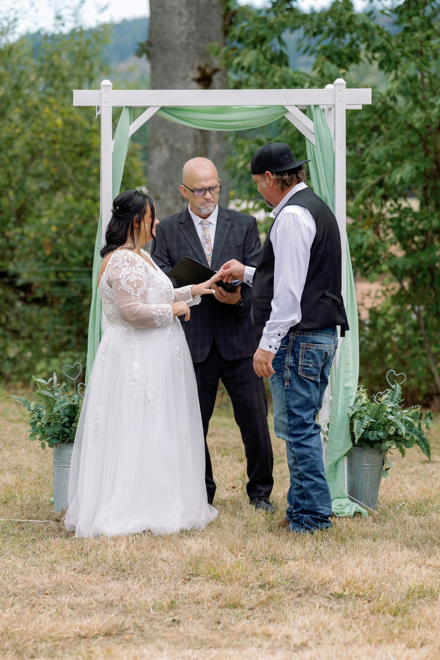 An intimate outdoor wedding ceremony takes place under a white arbor surrounded by greenery and trees.
