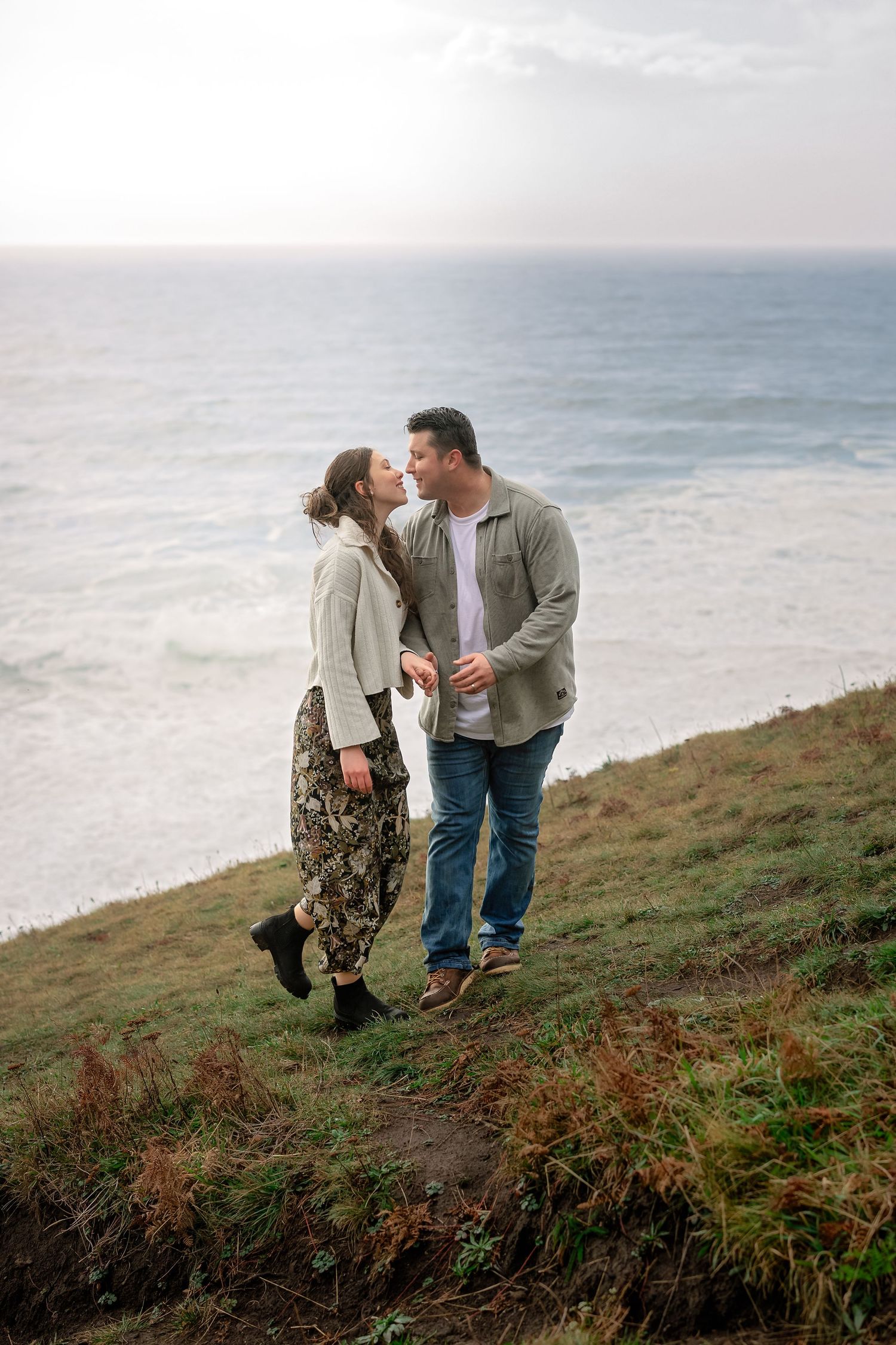 Two people walk along a misty coastal path with crashing waves visible in the distance.