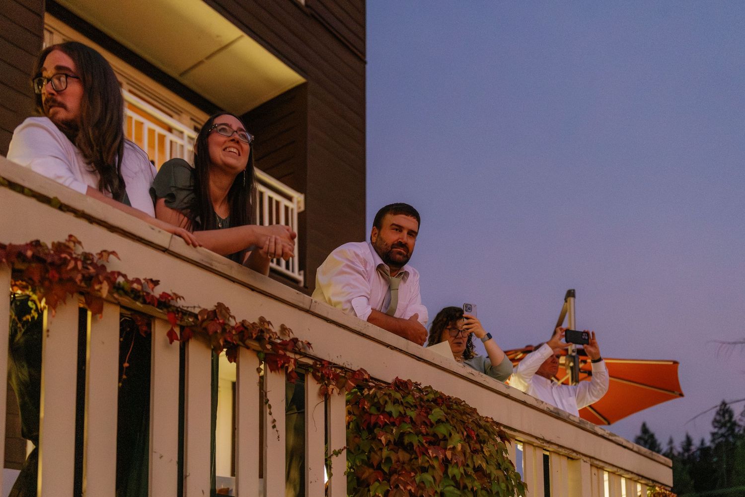 People gathered on a porch balcony enjoying drinks and conversation during an outdoor social gathering.