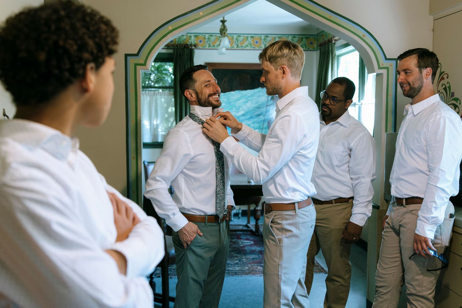 Groomsmen helping adjust each other's ties while getting ready in a room with an arched doorway.