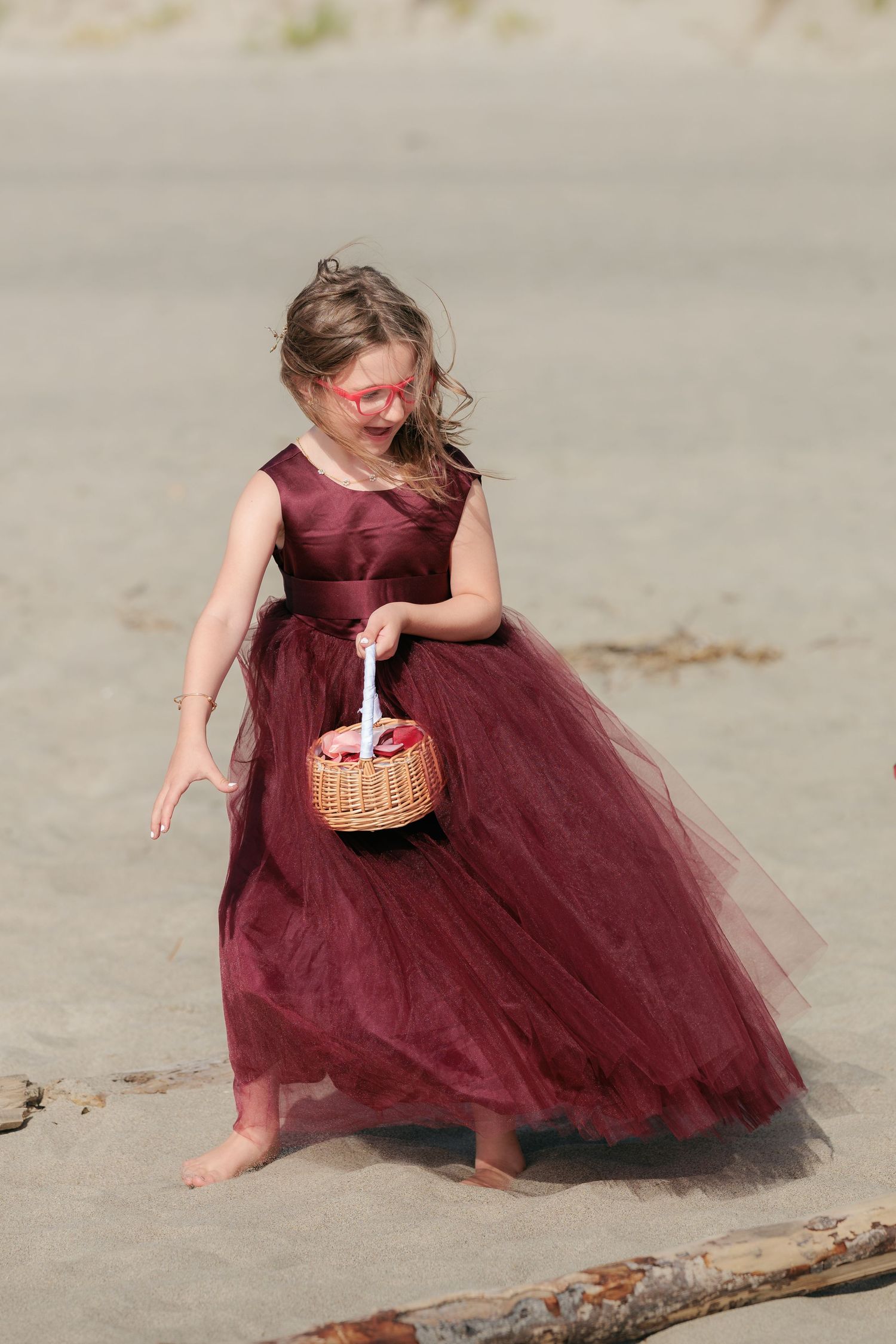 Young flower girl in a burgundy tulle dress carrying a basket on the beach.