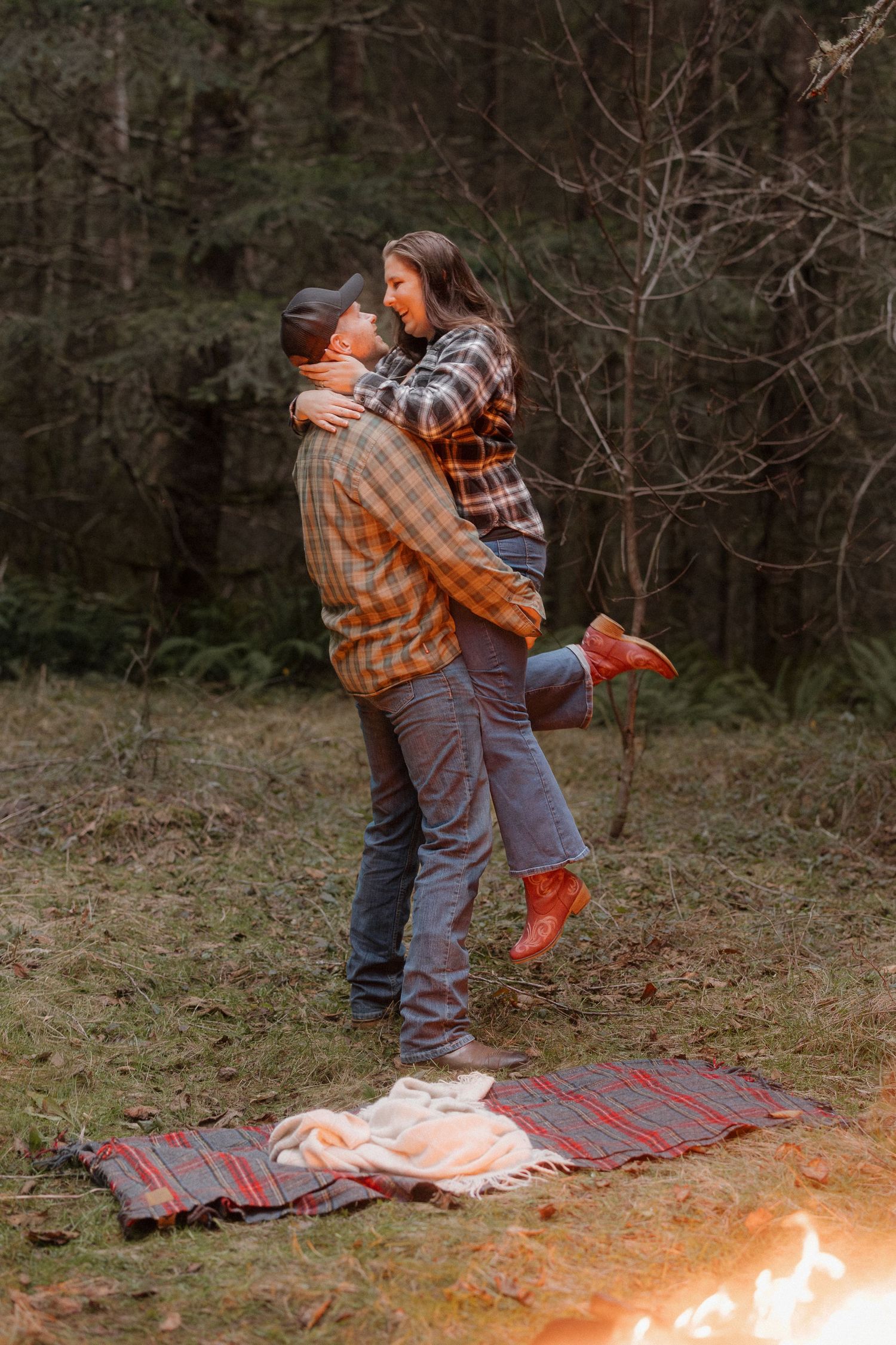 A couple dances and embraces on a plaid blanket near a glowing campfire in a wooded outdoor setting at dusk.