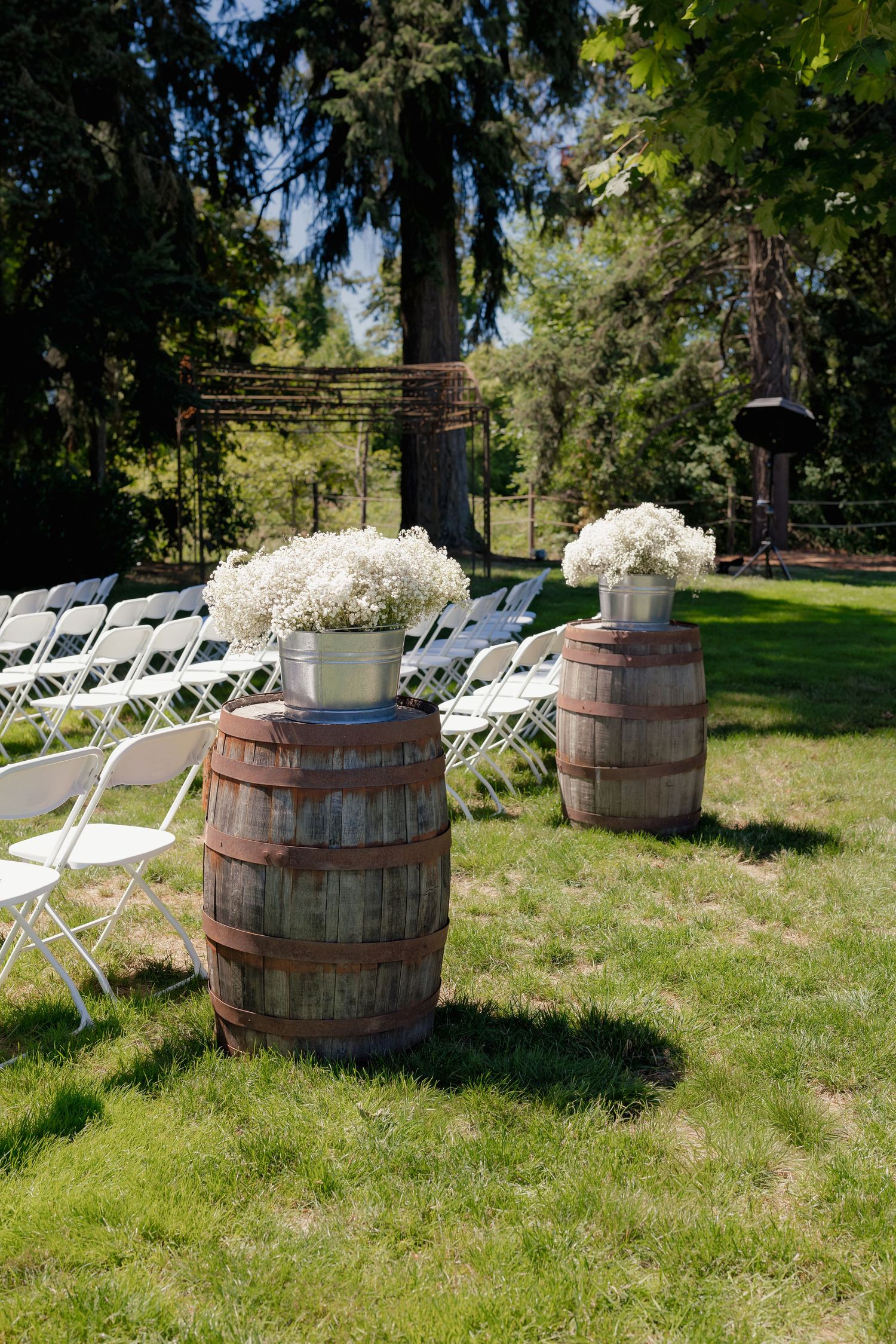 Rustic outdoor wedding ceremony setup with wooden barrels topped with white baby's breath flowers flanking white folding chairs.