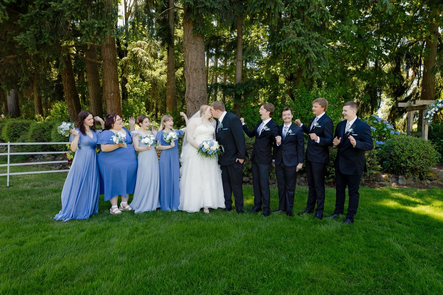 Wedding party poses outdoors, bridesmaids in blue gowns and groomsmen in suits, as bride and groom share a kiss on green lawn.