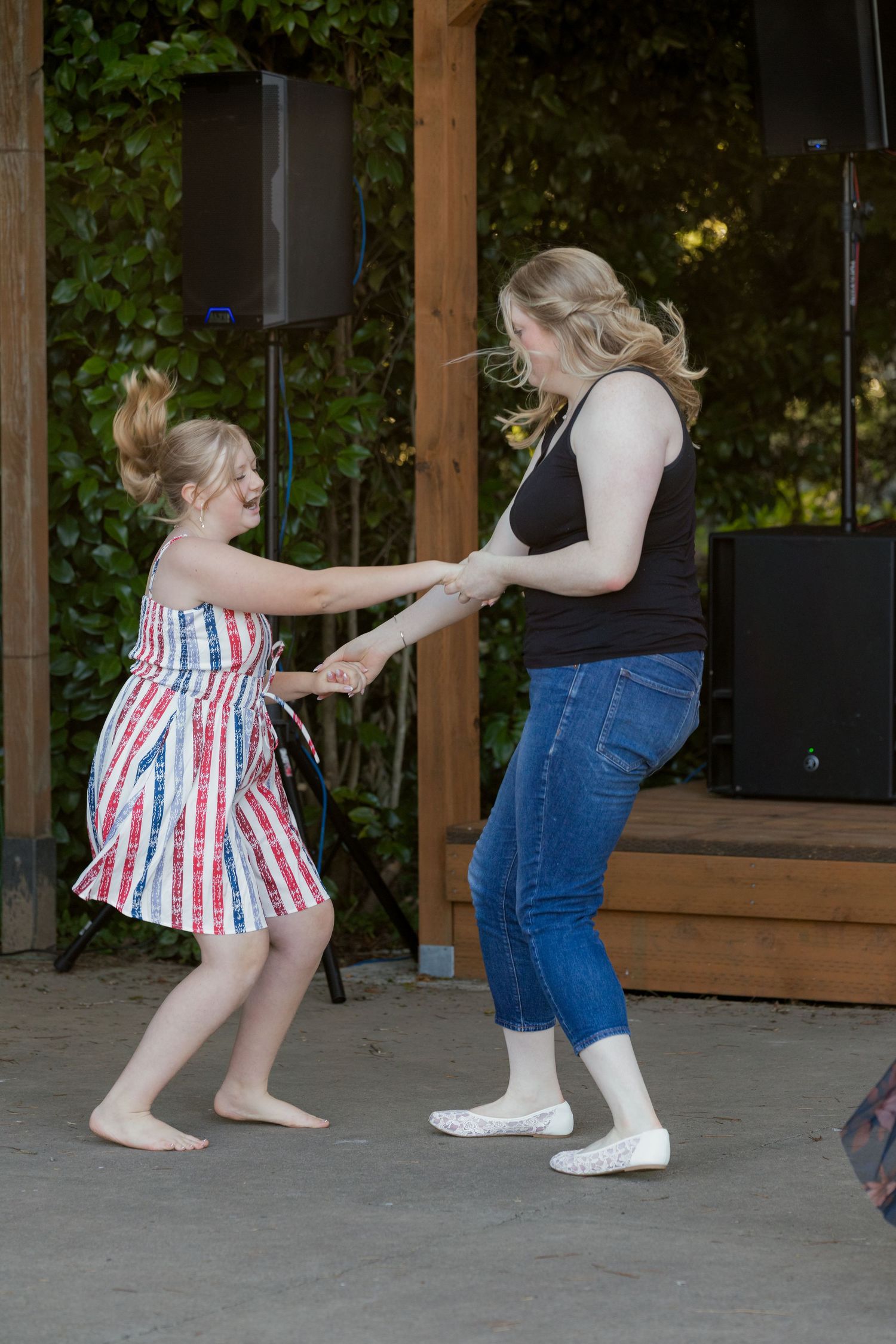 Young girl in striped dress dancing and holding hands with a woman in jeans at an outdoor wedding reception.