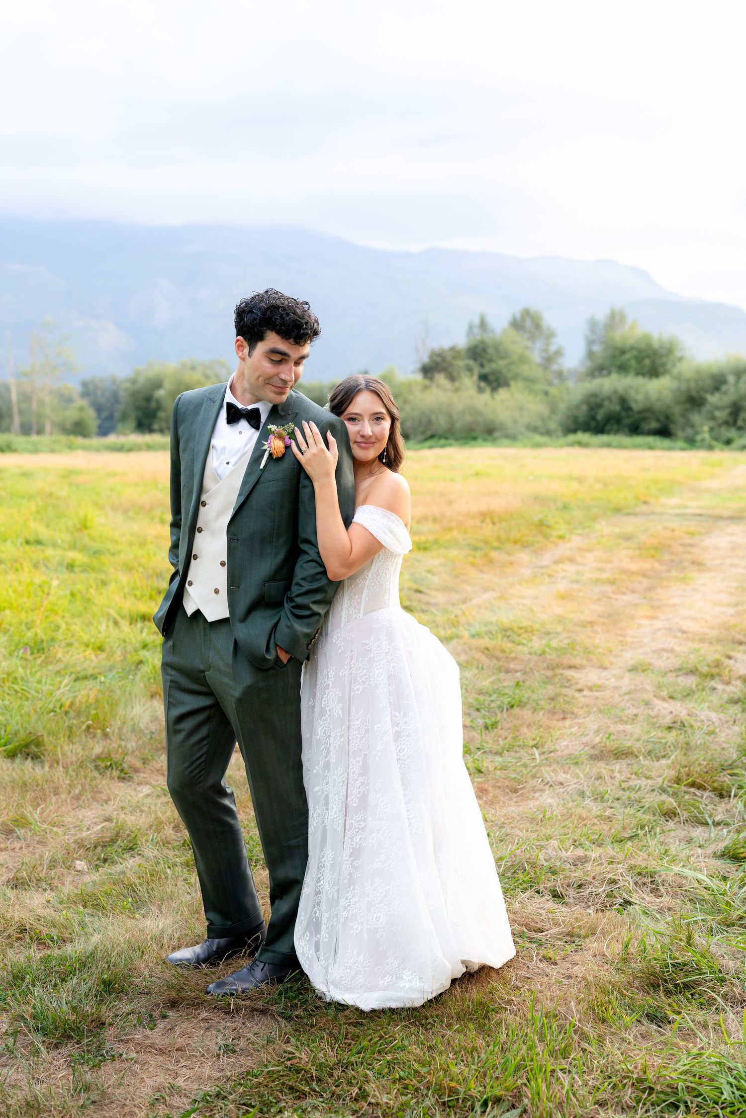 Bride in off-shoulder white gown leans on groom in dark green tuxedo in a scenic mountain meadow.