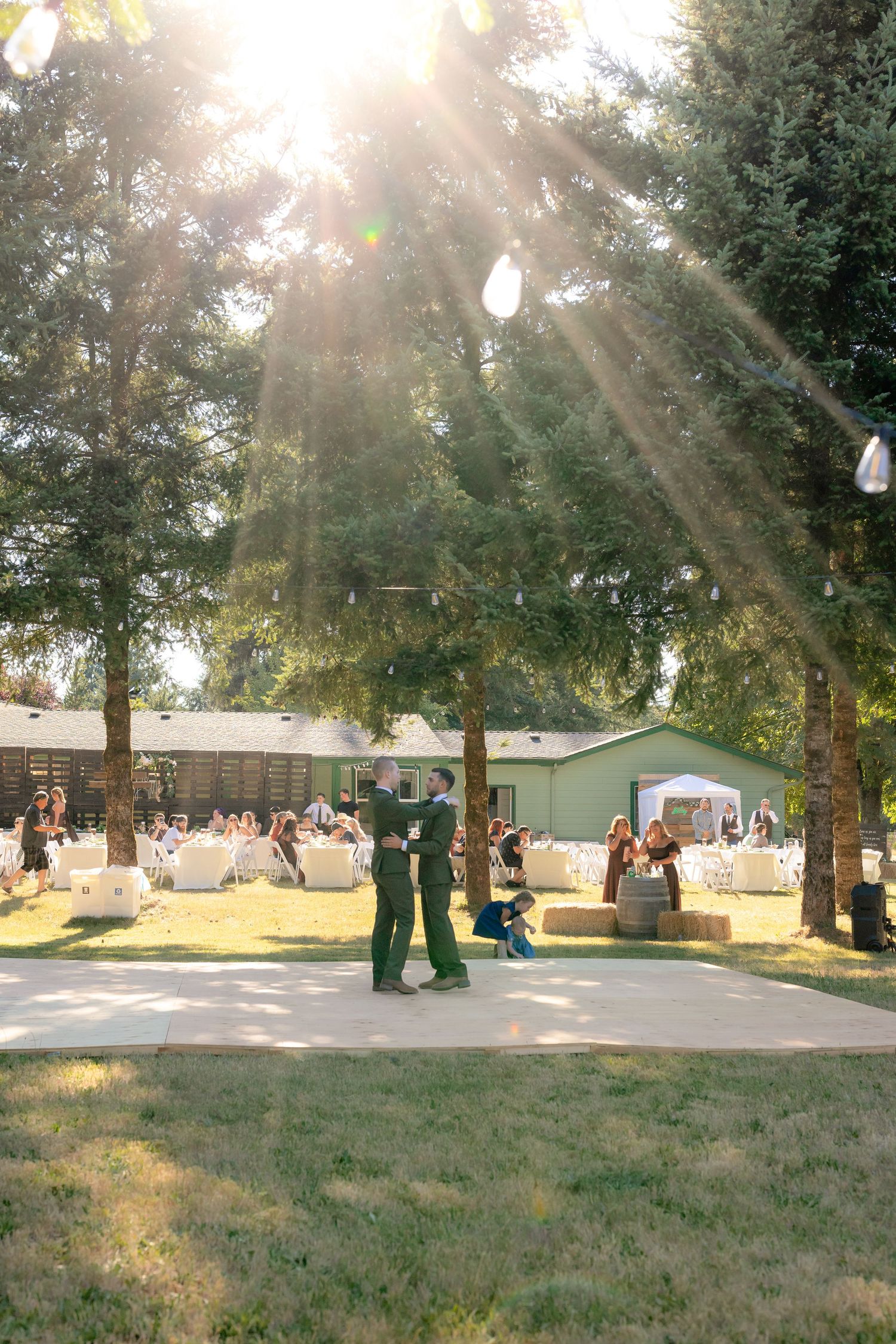 Sunlit outdoor wedding reception with guests seated among tall trees, two people dancing in the foreground.
