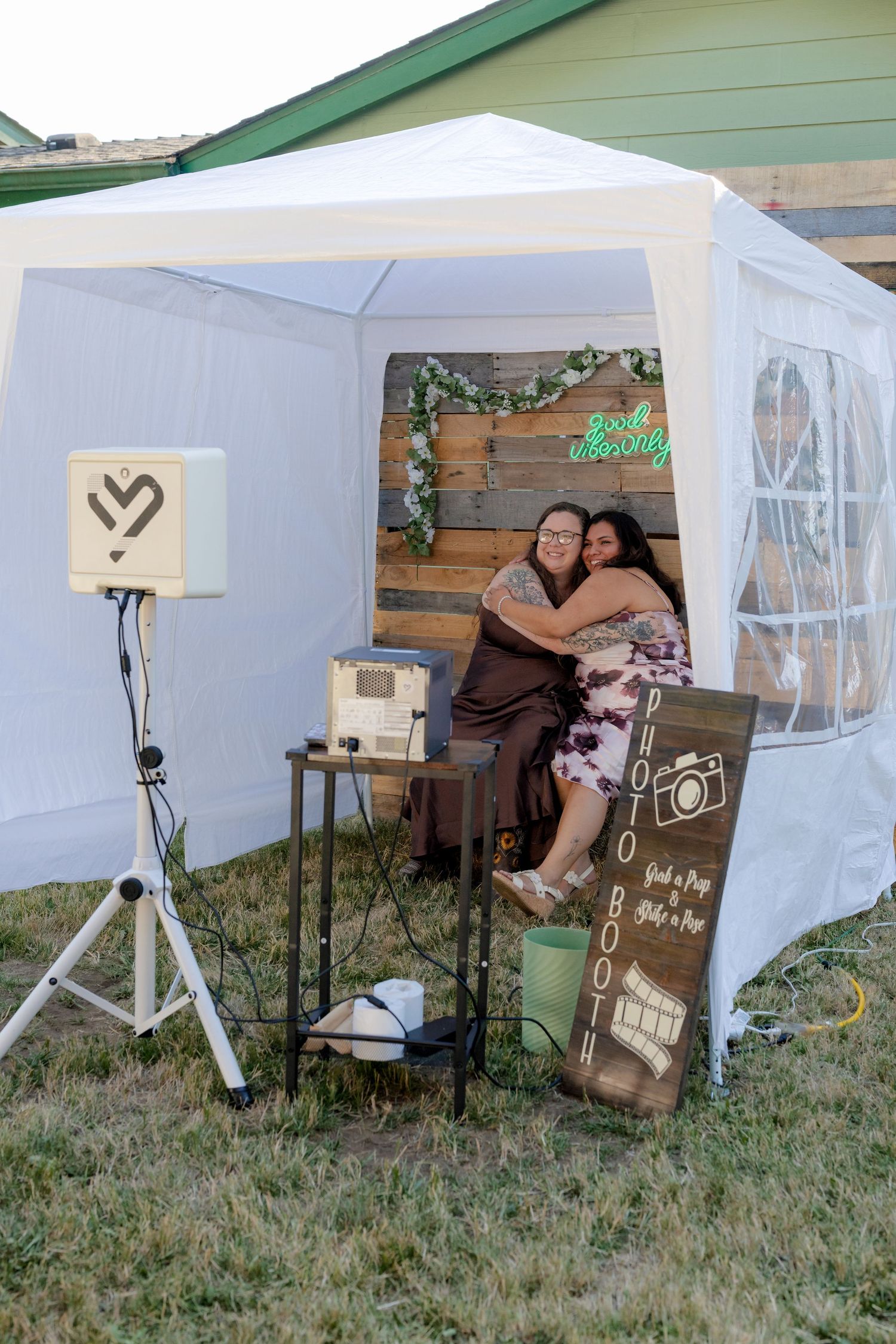 Two women embrace in a rustic photo booth setup at an outdoor wedding reception.