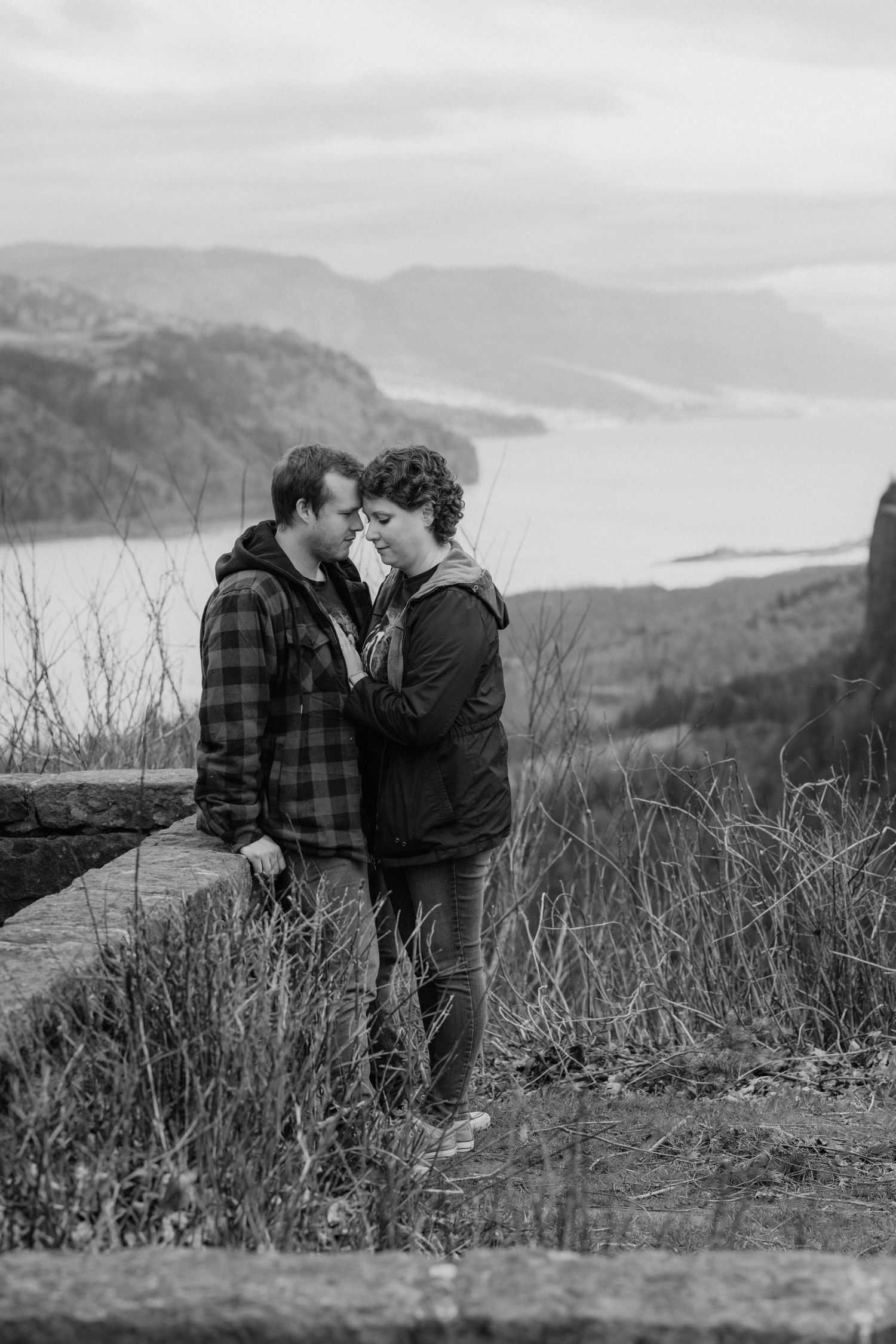 A couple embraces on a scenic cliff overlooking mountains and water in black and white photography.