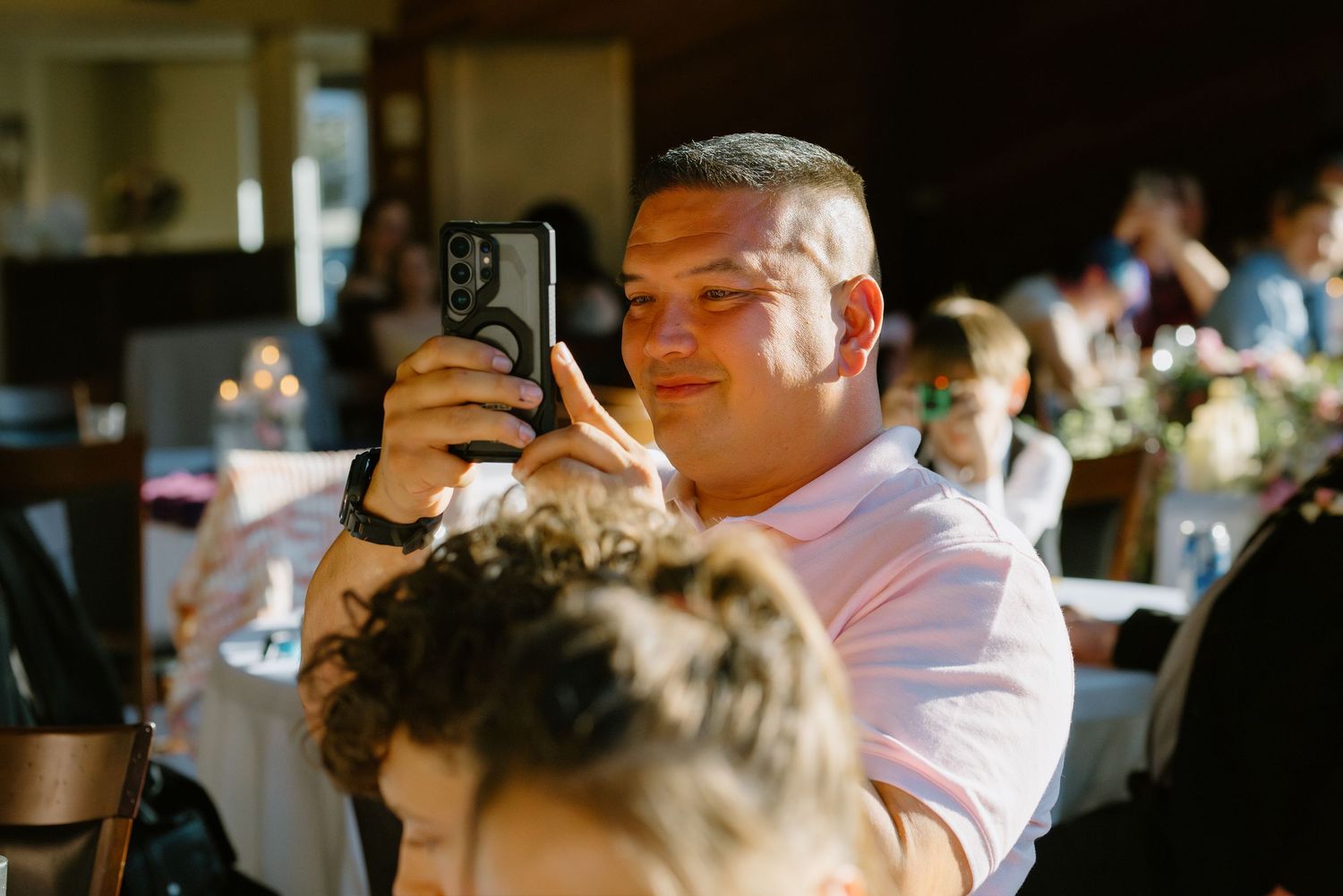 Male wedding guest in pink shirt smiling while taking a photo with his smartphone at a reception.
