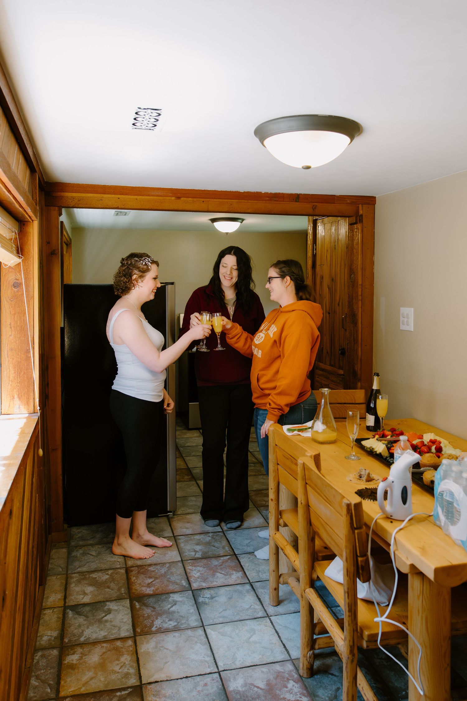 Three women toasting with wine glasses in a cozy kitchen near a wooden dining table with food and drinks.