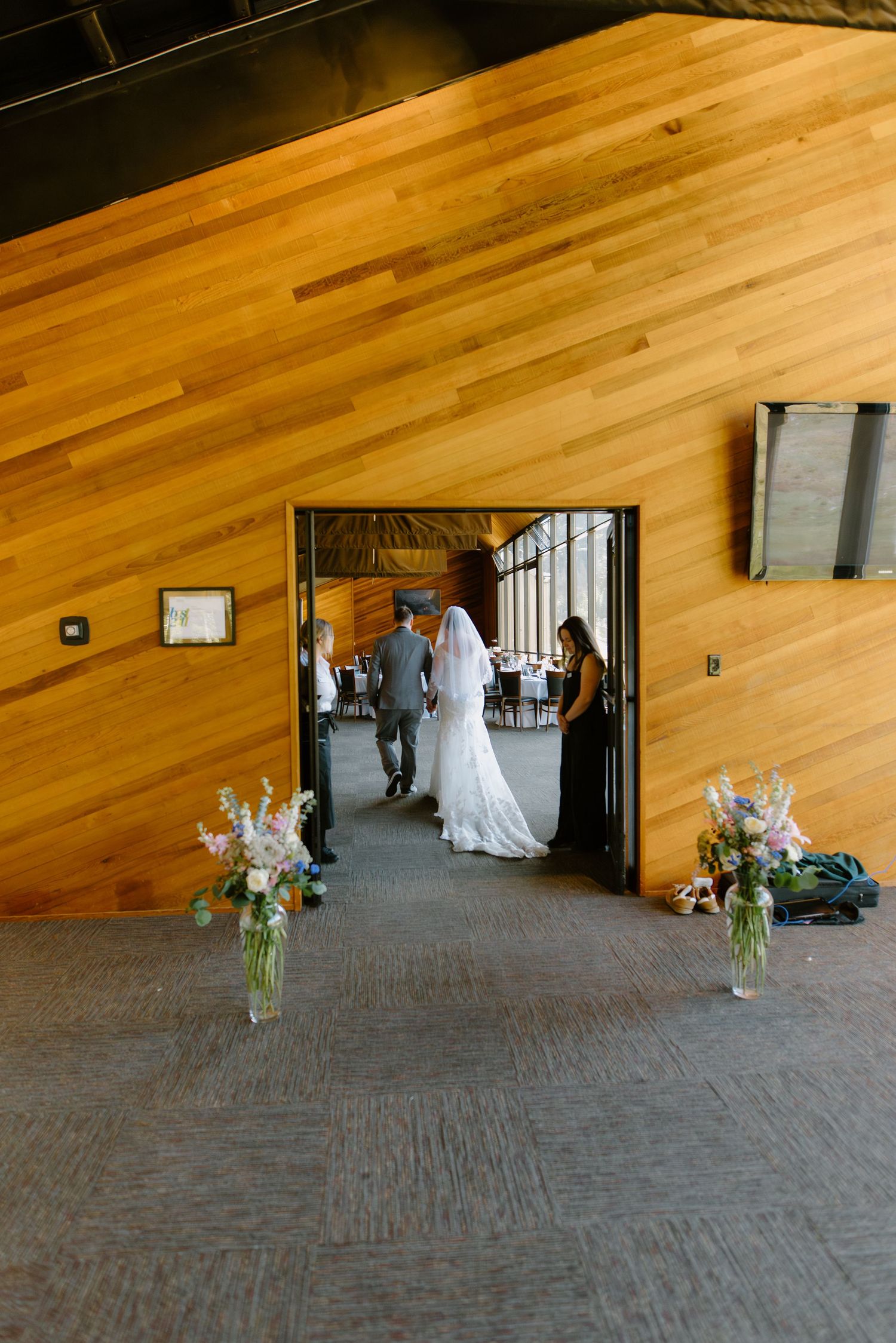Bride and groom enter wedding reception venue through wooden doors flanked by floral arrangements on carpet.