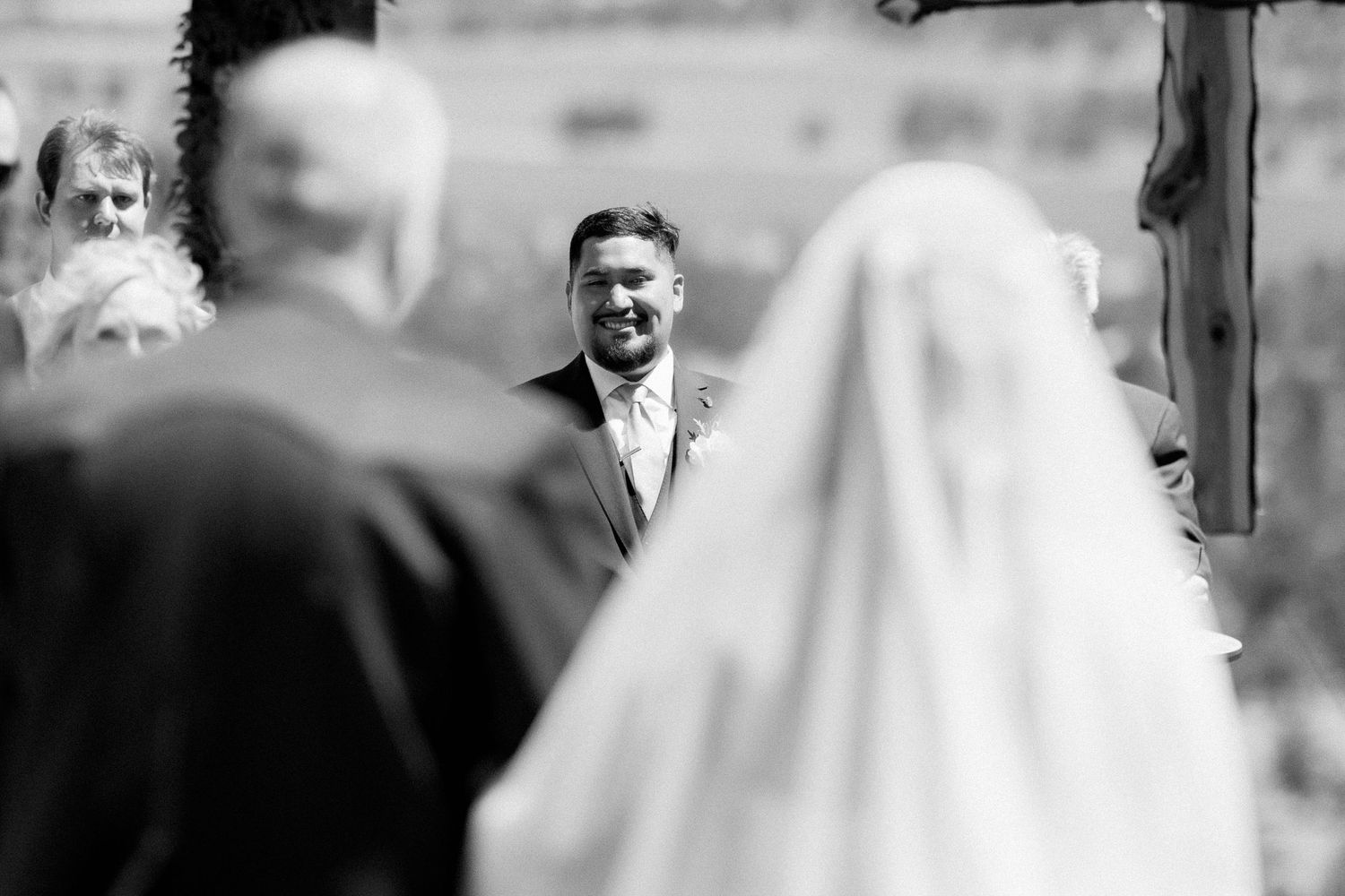Groom smiling as bride walks down the aisle at outdoor wedding ceremony, captured in black and white.
