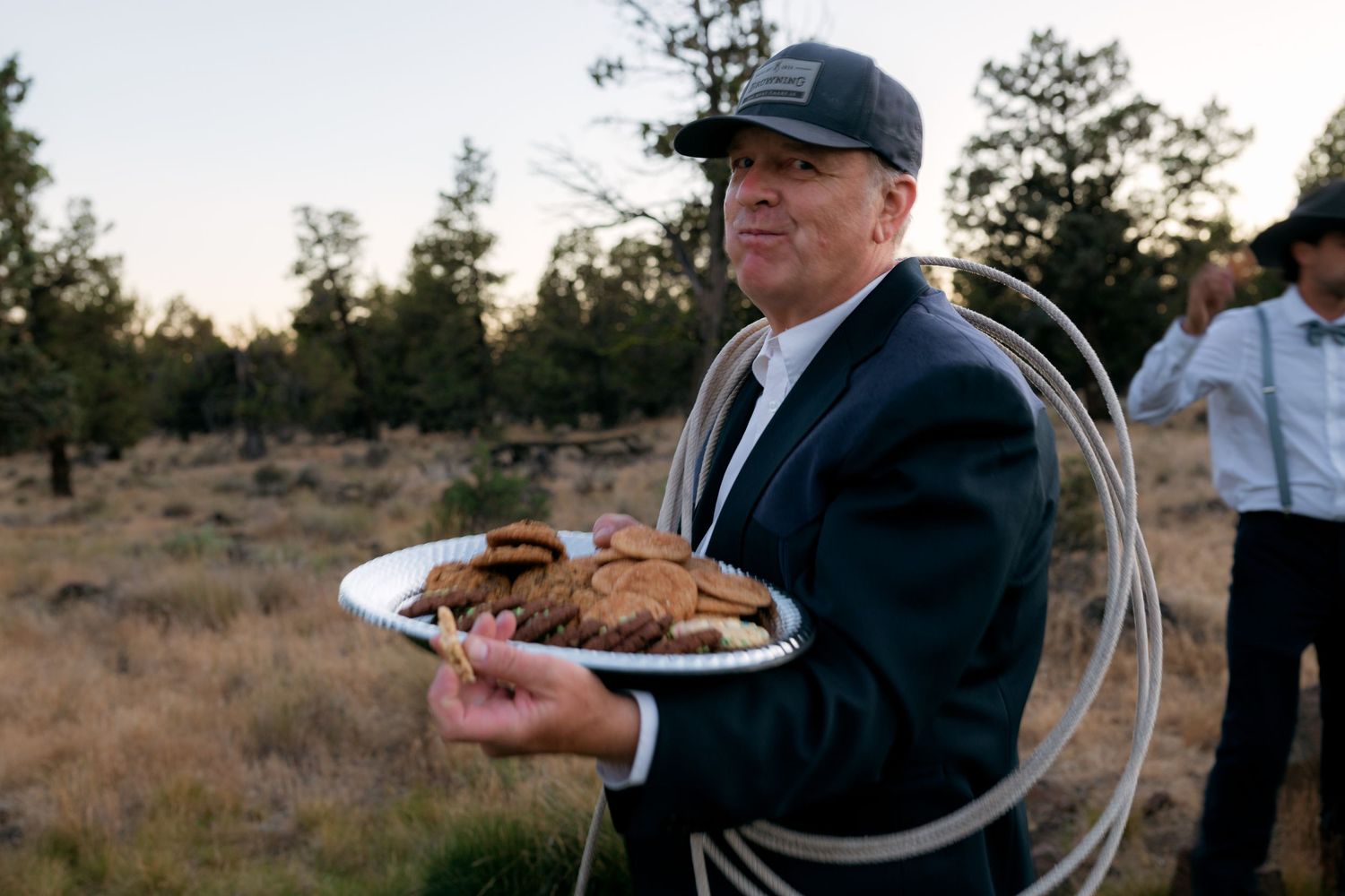 Man in suit jacket holds plate of cookies outdoors at twilight during wedding celebration.