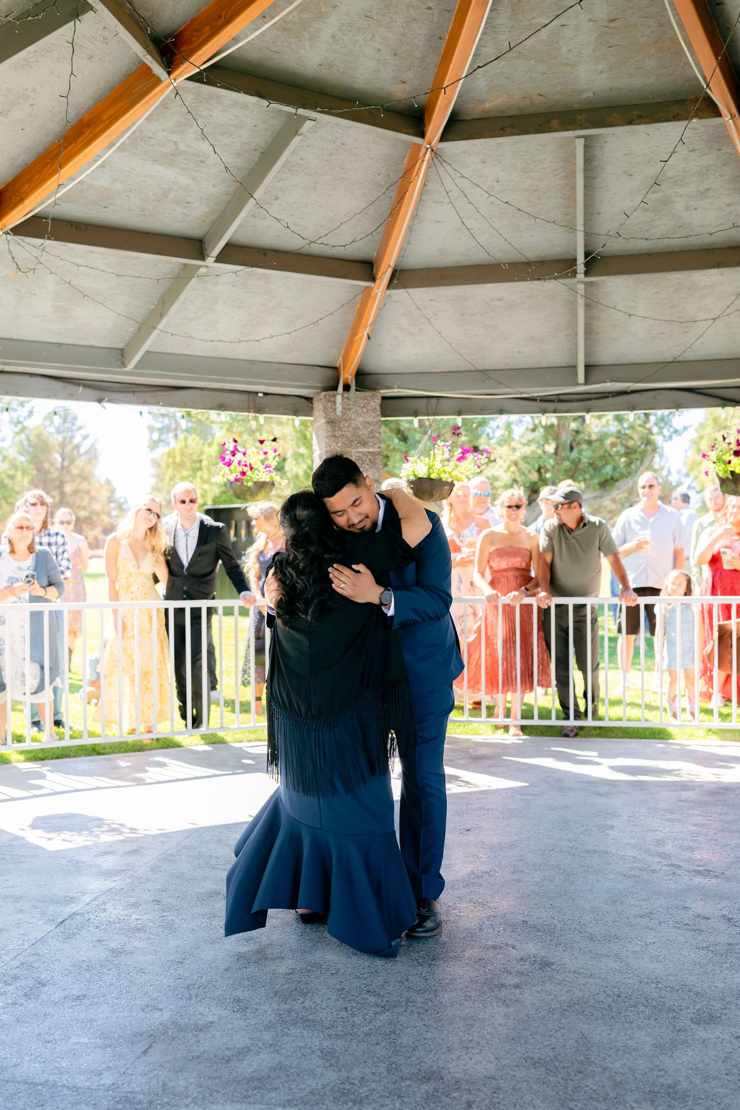 Couple shares emotional first dance under a pavilion surrounded by wedding guests outdoors.