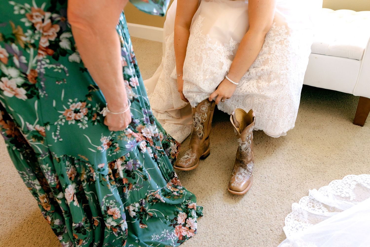 Bride in lace wedding dress putting on embroidered cowboy boots assisted by bridesmaid in green floral dress.