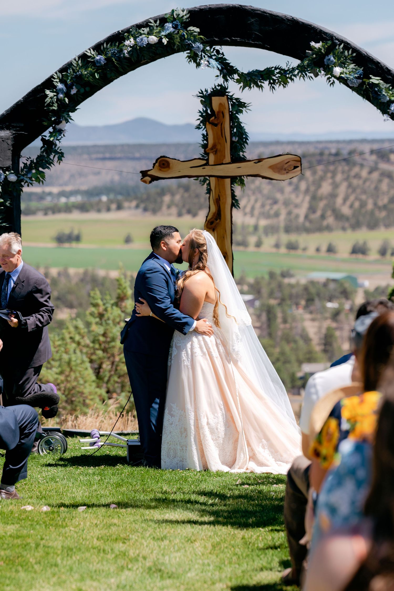 Bride and groom share first kiss under floral arch with wooden cross at outdoor wedding ceremony.