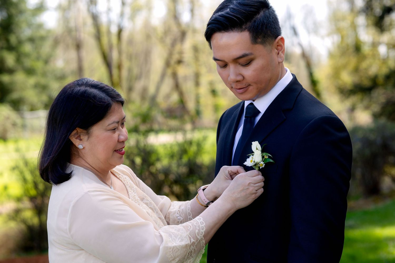 Older woman pinning a boutonniere on a young man in a black suit at an outdoor wedding.