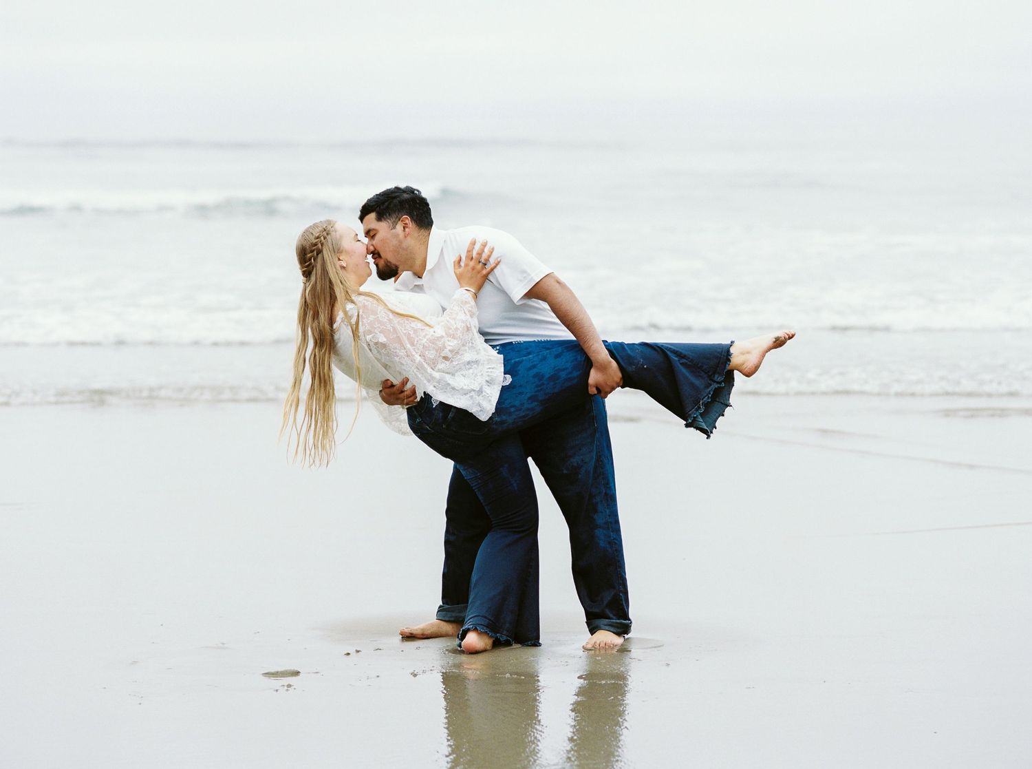 Couple sharing a romantic kiss on a sandy beach, man lifting woman while waves crash in the background.