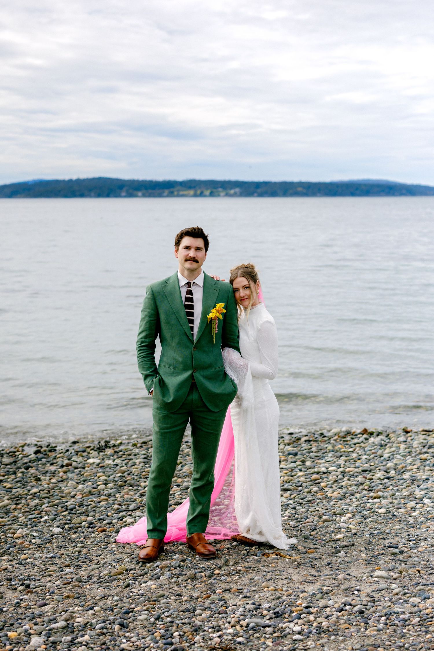 Wedding couple standing on pebble beach with calm ocean backdrop, groom in green suit and bride in white dress with pink train.