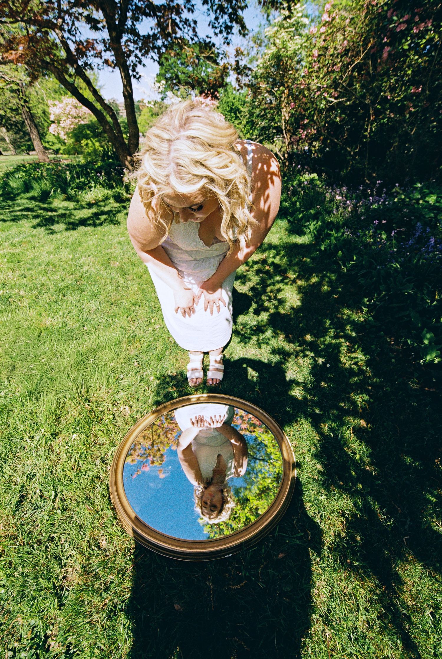 Blonde woman in white dress looking into a round gold mirror reflecting blue sky, standing on lush green grass outdoors.