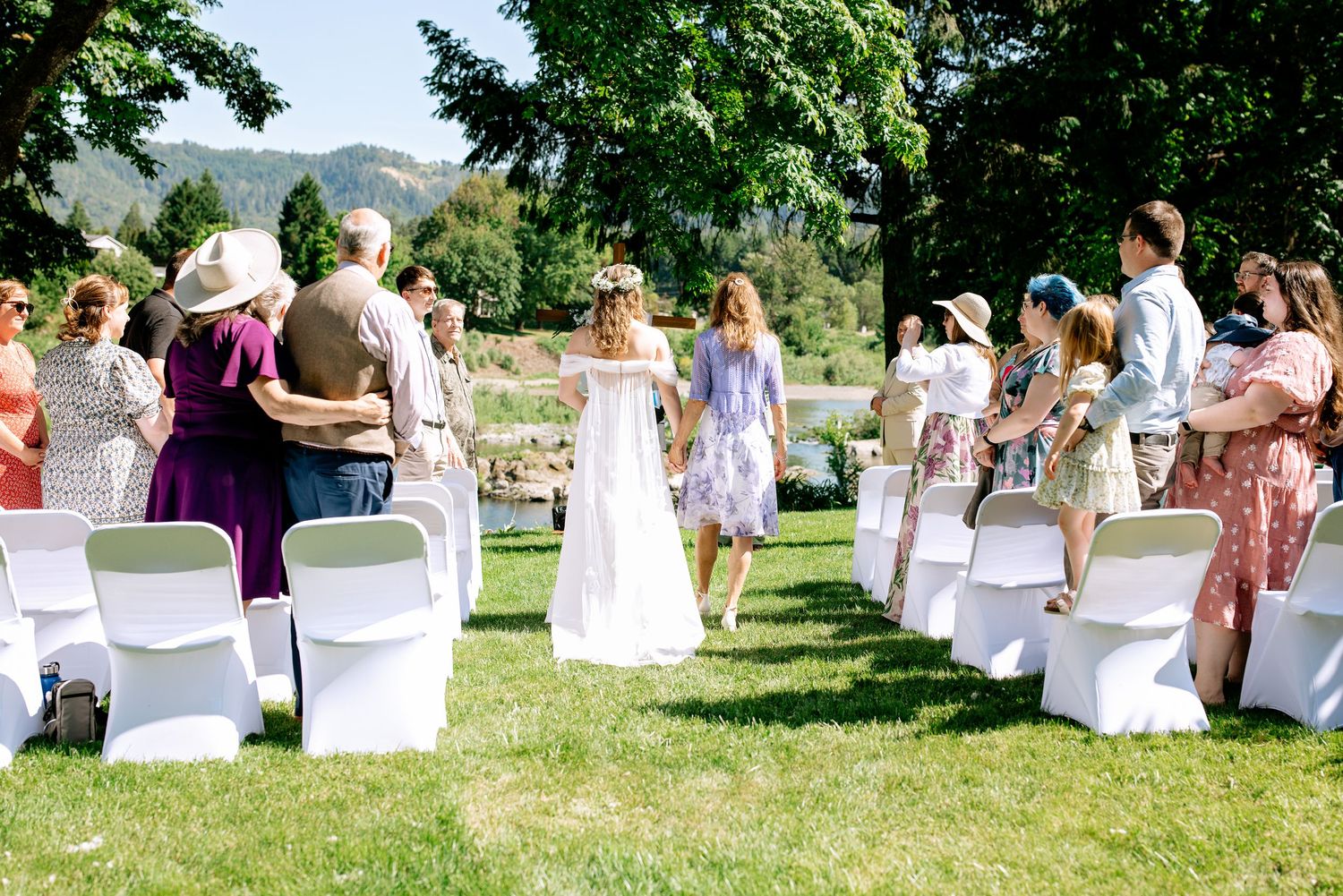Bride walks down outdoor wedding aisle on sunny day with white chairs and mountain backdrop.