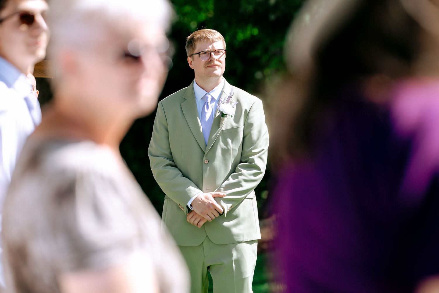 Groom in sage green suit standing attentively during outdoor wedding ceremony with guests in the foreground.