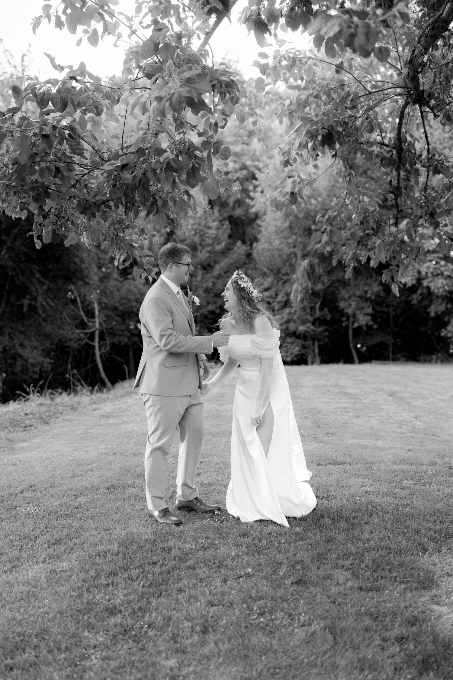 Black and white photo of bride and groom holding hands and dancing on grass beneath leafy trees outdoors.