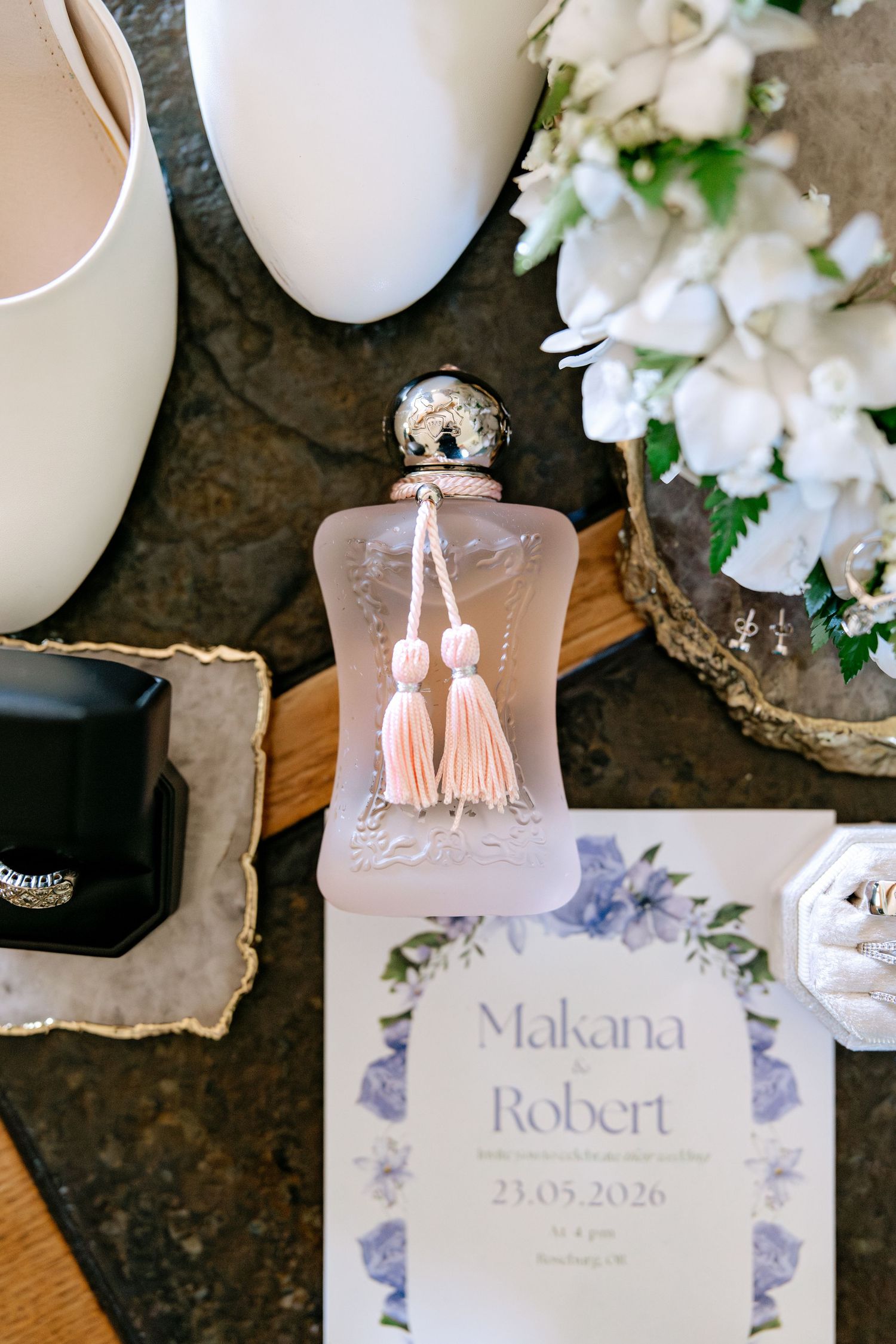 Wedding flat lay with perfume bottle, invitation for Makana and Robert dated 23.05.2026, bridal shoes, and white flowers.
