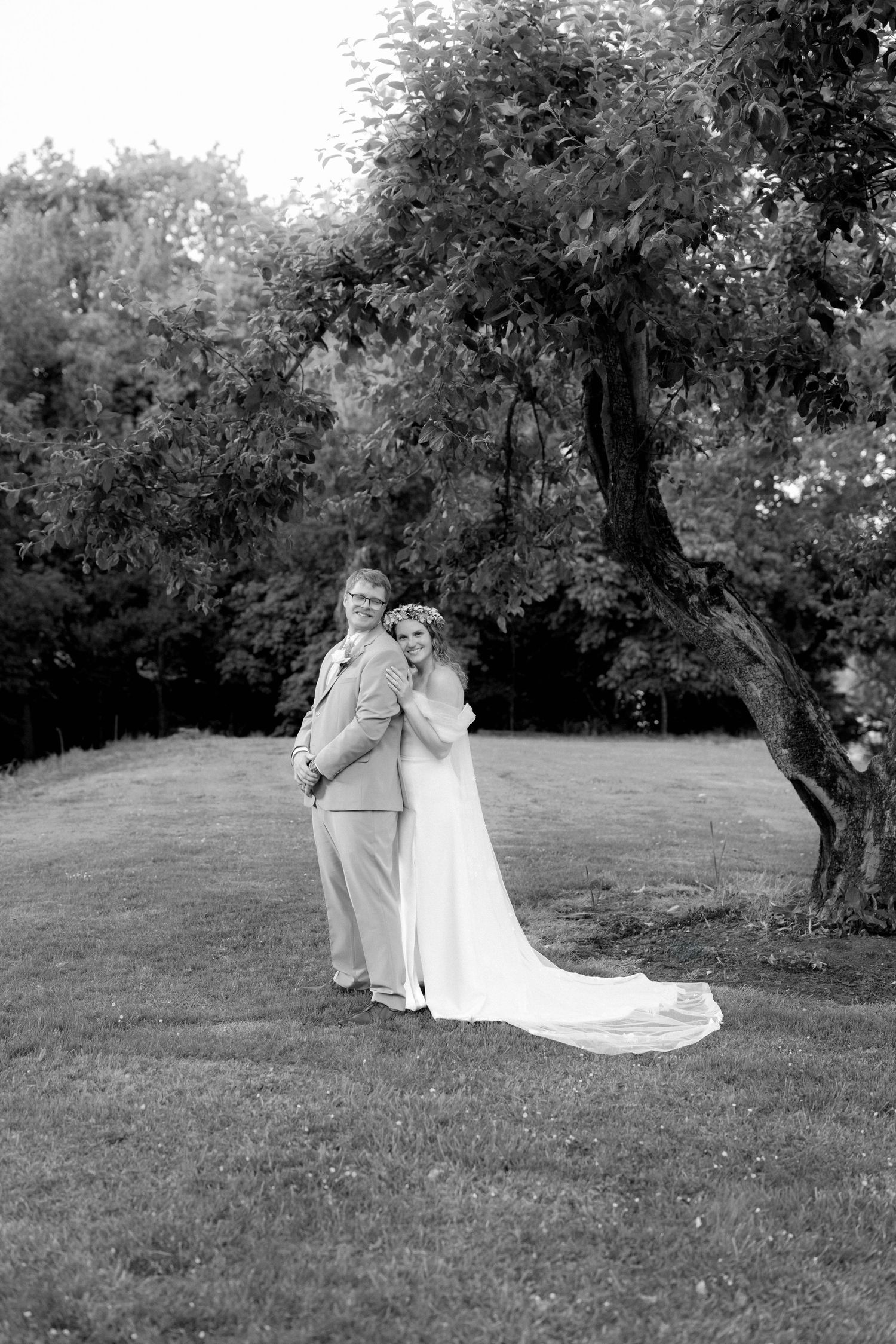 Black and white photo of a bride and groom embracing outdoors near a large tree on their wedding day.