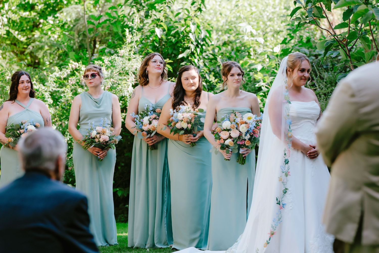 Bridesmaids in sage green dresses holding bouquets stand outdoors beside a bride in white at a garden wedding.
