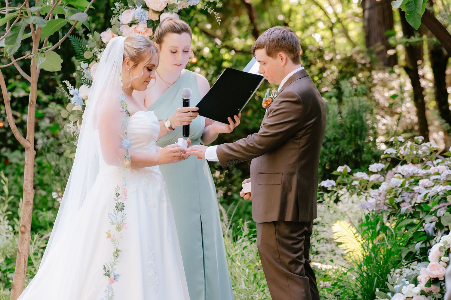 Bride and groom exchange rings during outdoor wedding ceremony with officiant holding black book amid lush garden greenery.