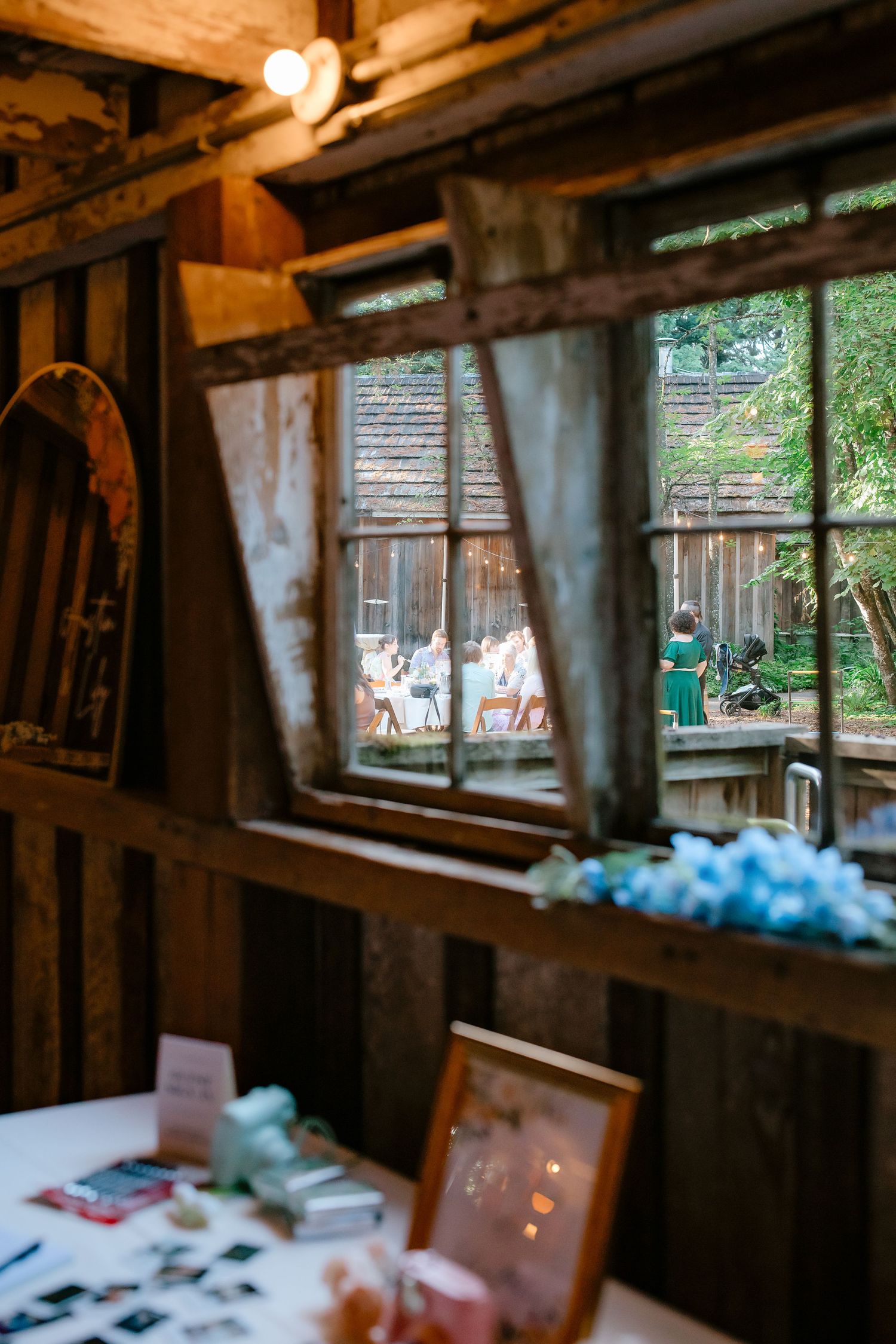 Rustic barn wedding venue interior with wooden beams and window view of outdoor reception guests.