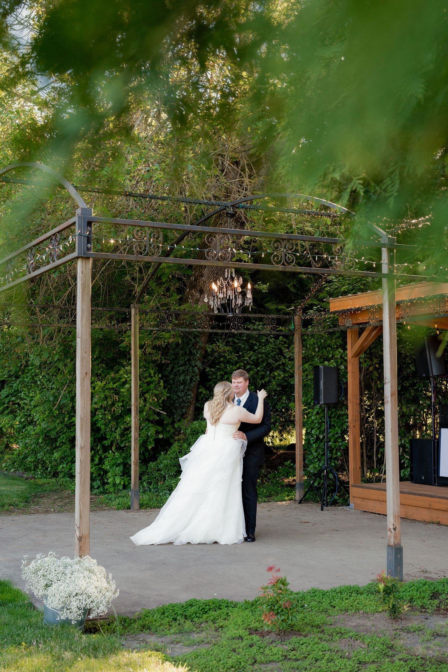 Bride and groom share their first dance under an elegant iron pergola with chandelier surrounded by lush greenery.