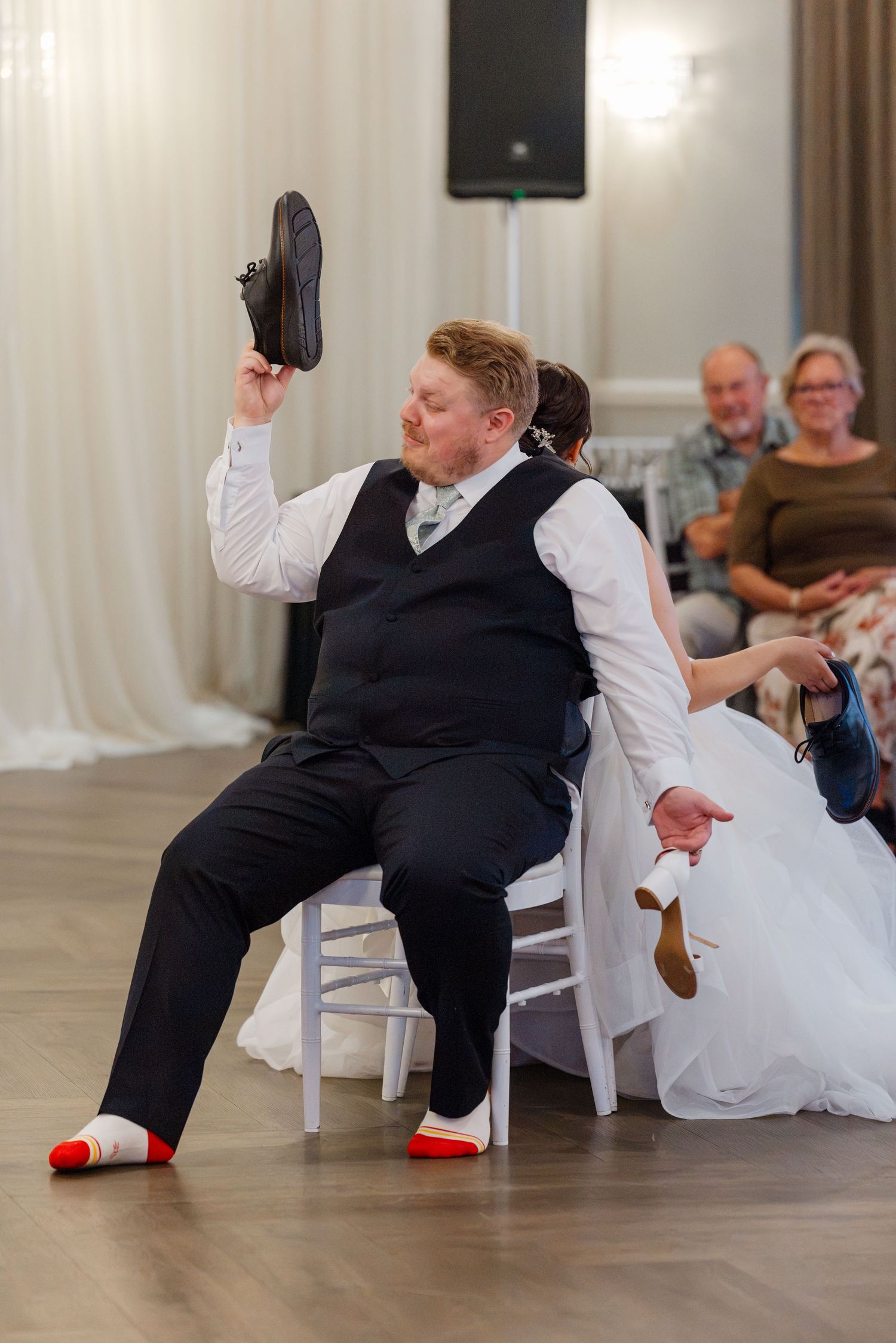Groom playfully holds up shoe during wedding reception shoe game while bride laughs behind him.