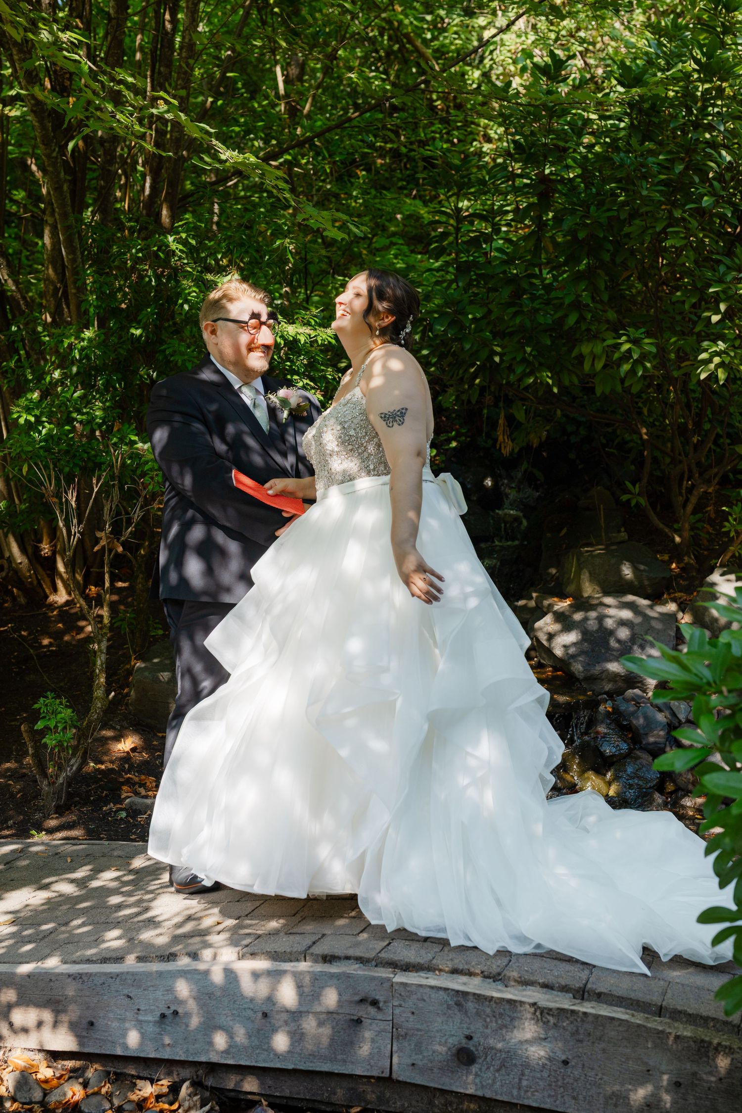 Bride and groom share a joyful moment on a garden bridge, bride in layered white gown with tattoos visible.