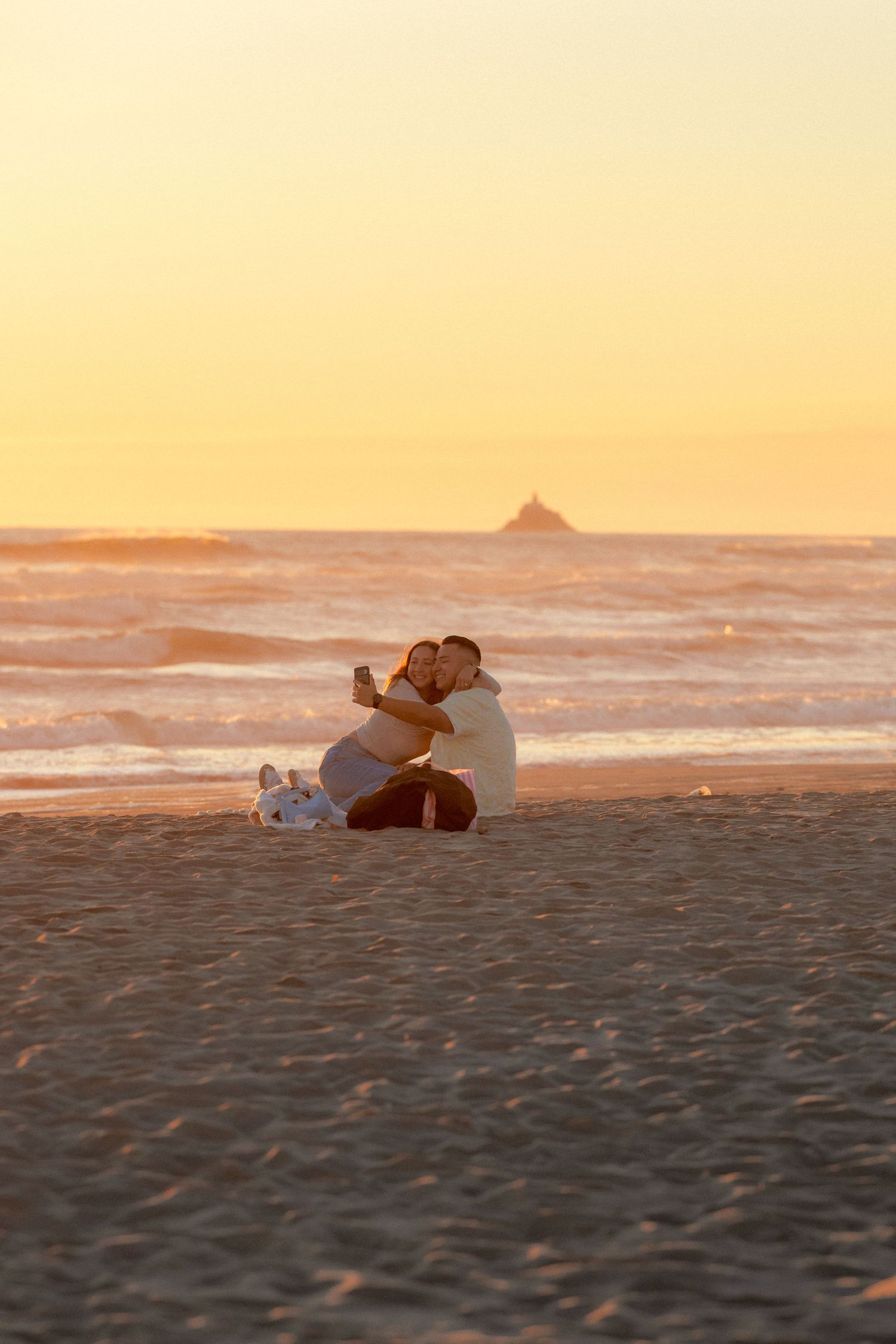 Couple sitting together on beach taking selfie during vibrant orange sunset by the ocean.