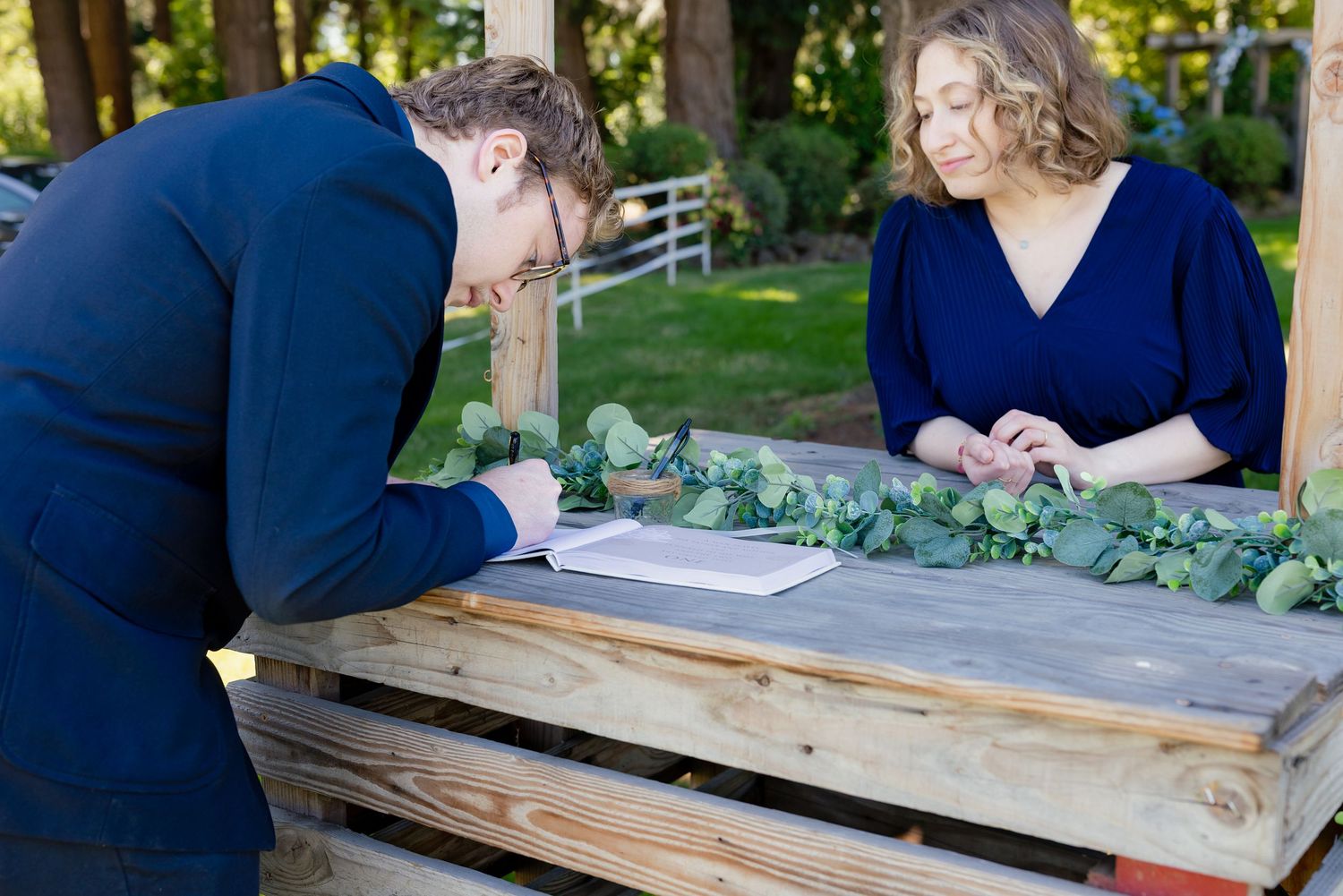 Man in navy suit signing documents at wooden table with woman in blue dress during outdoor wedding ceremony.