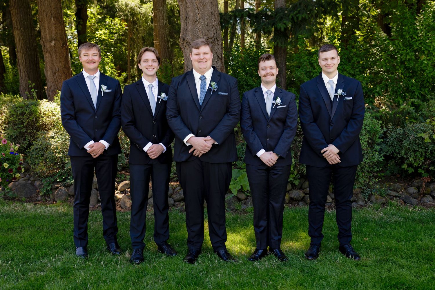 Five groomsmen in matching dark suits with boutonnieres pose together on a green lawn outdoors.