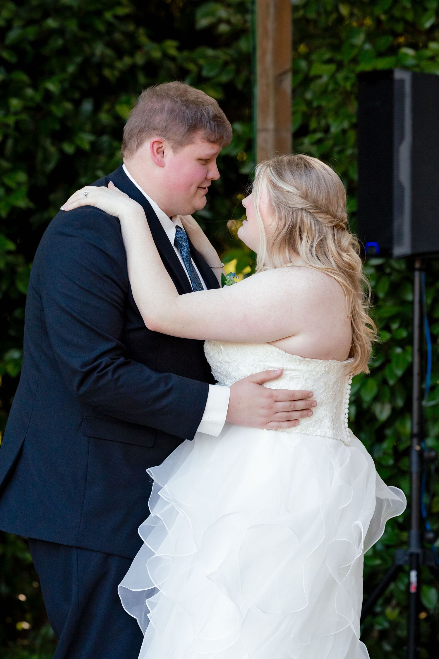 Bride and groom sharing their first dance outdoors, embracing closely with green hedge backdrop and speaker visible.