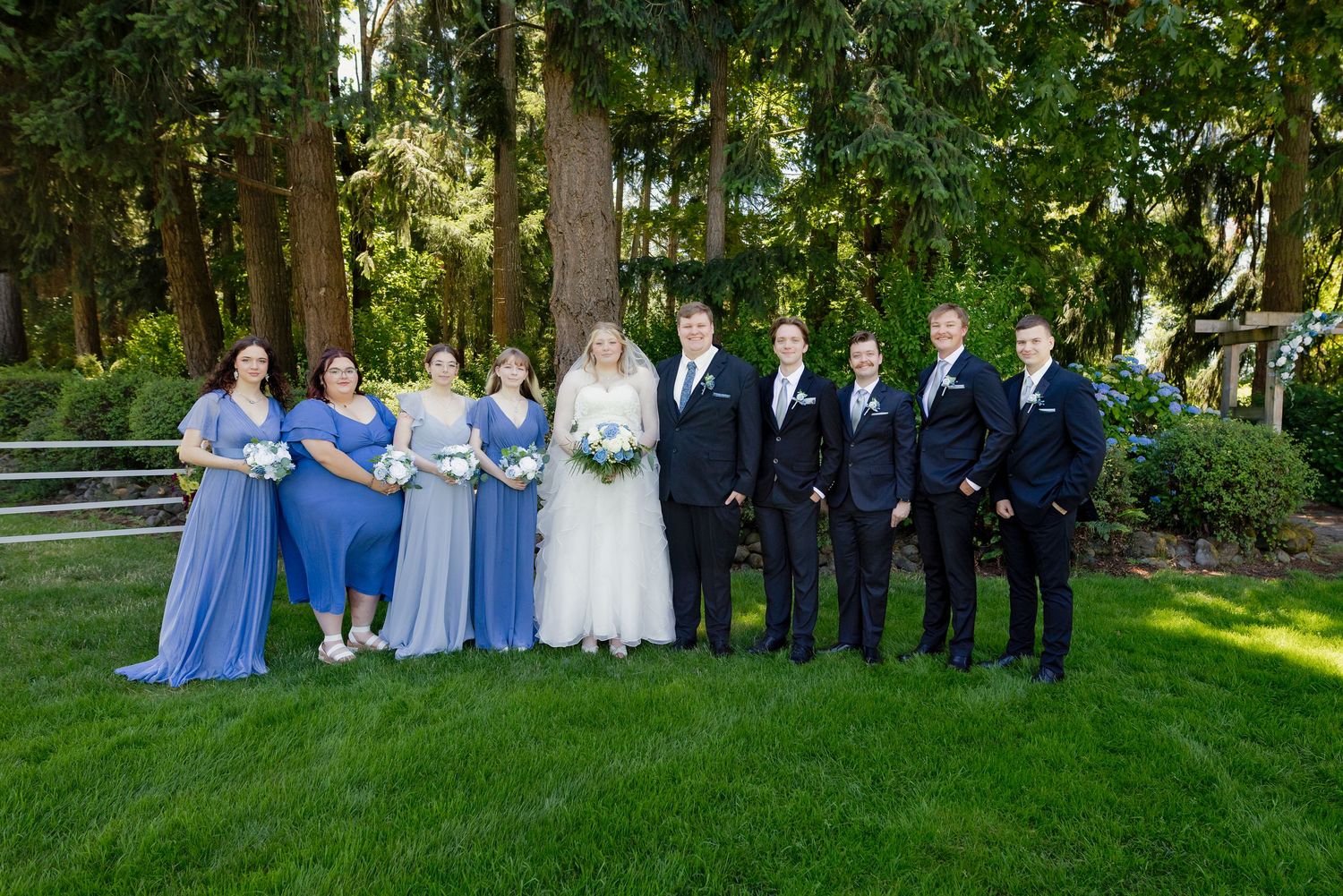 Wedding party posing on green lawn with bridesmaids in blue dresses and groomsmen in dark suits.