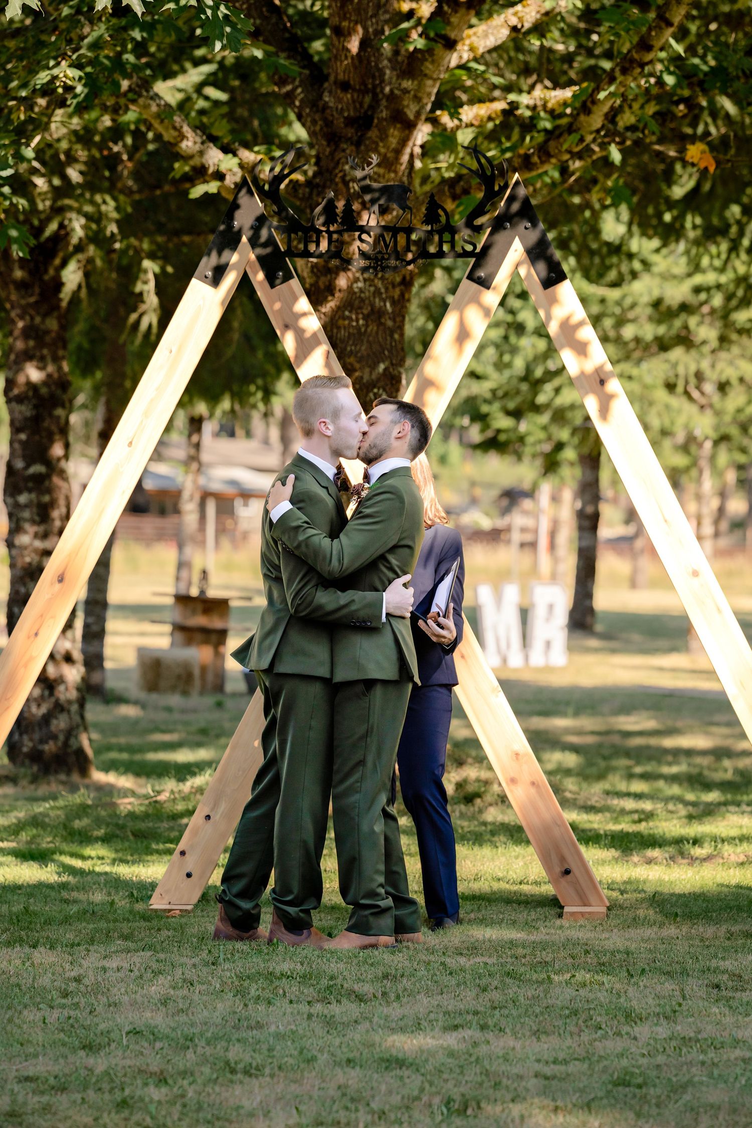 Two grooms in matching green suits share a first kiss under a wooden triangular arch at their outdoor wedding.