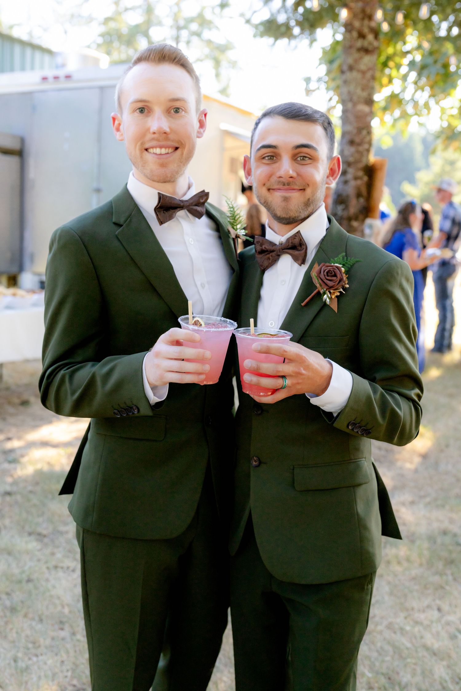 Two grooms in matching dark green suits with bow ties holding pink drinks at their outdoor wedding celebration.