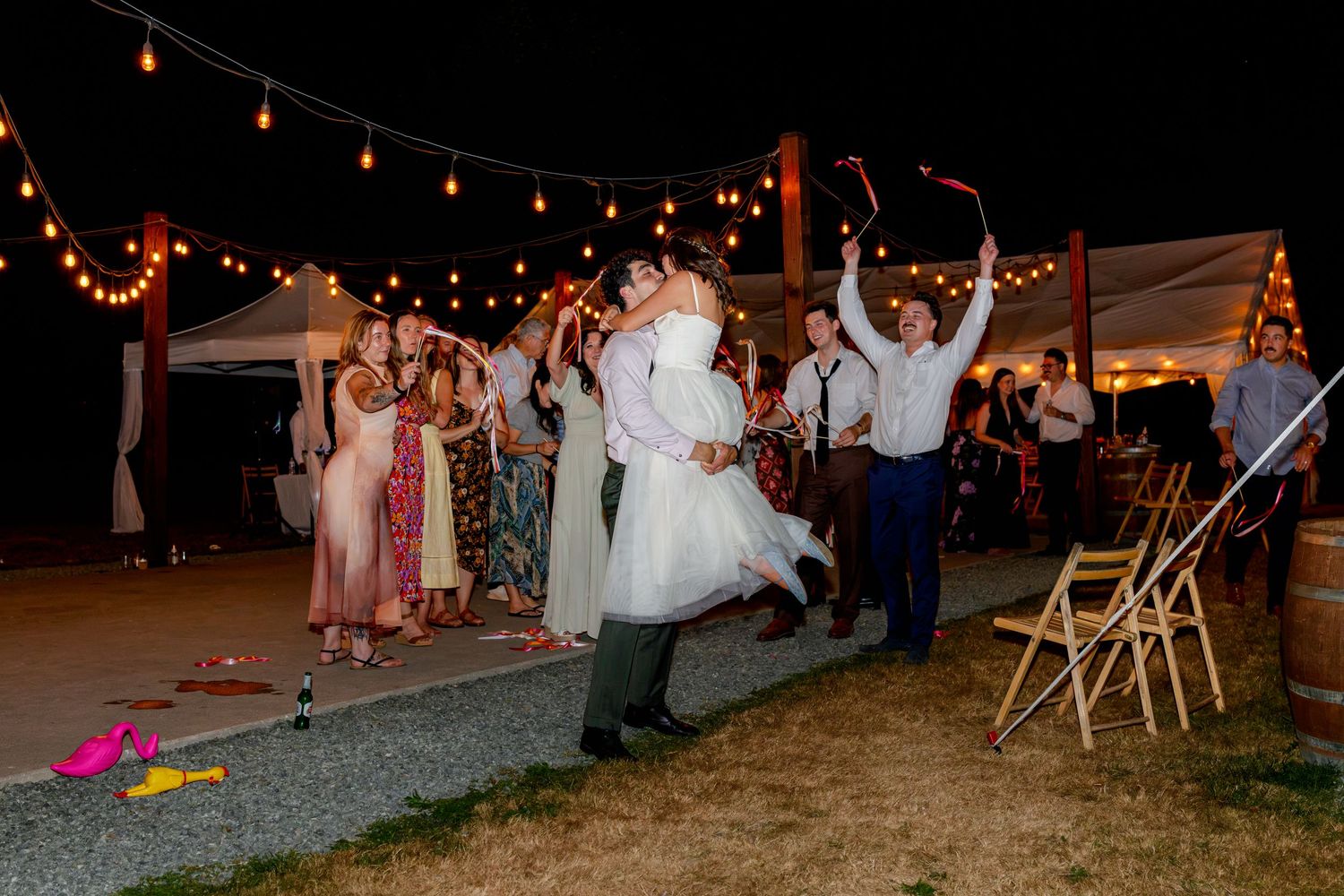 Bride and groom share first dance kiss outdoors at night wedding reception with string lights and cheering guests.