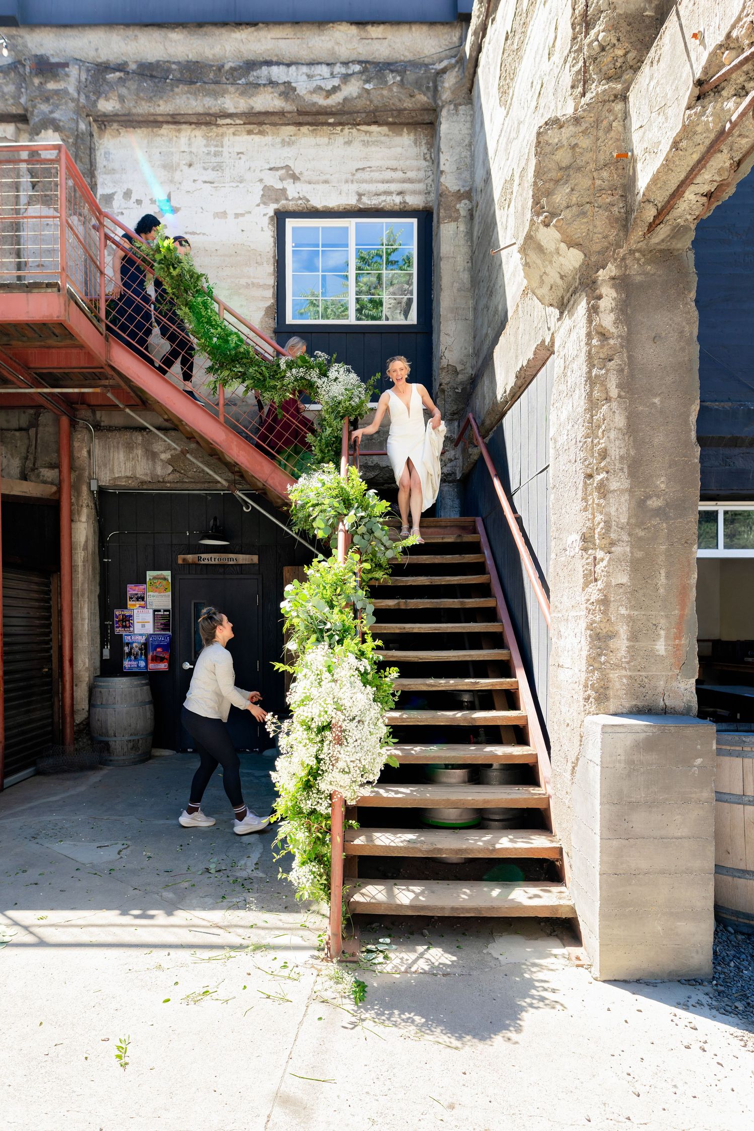Bride in white dress descends flower-adorned wooden staircase at rustic industrial wedding venue.