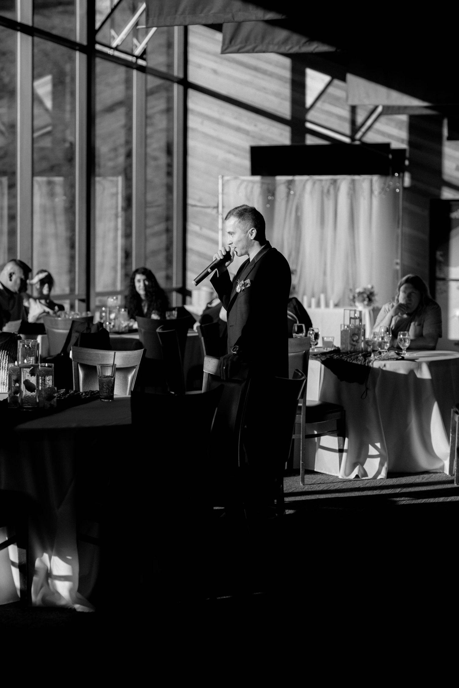 Black and white photo of man giving a speech with microphone at an elegant wedding reception venue.