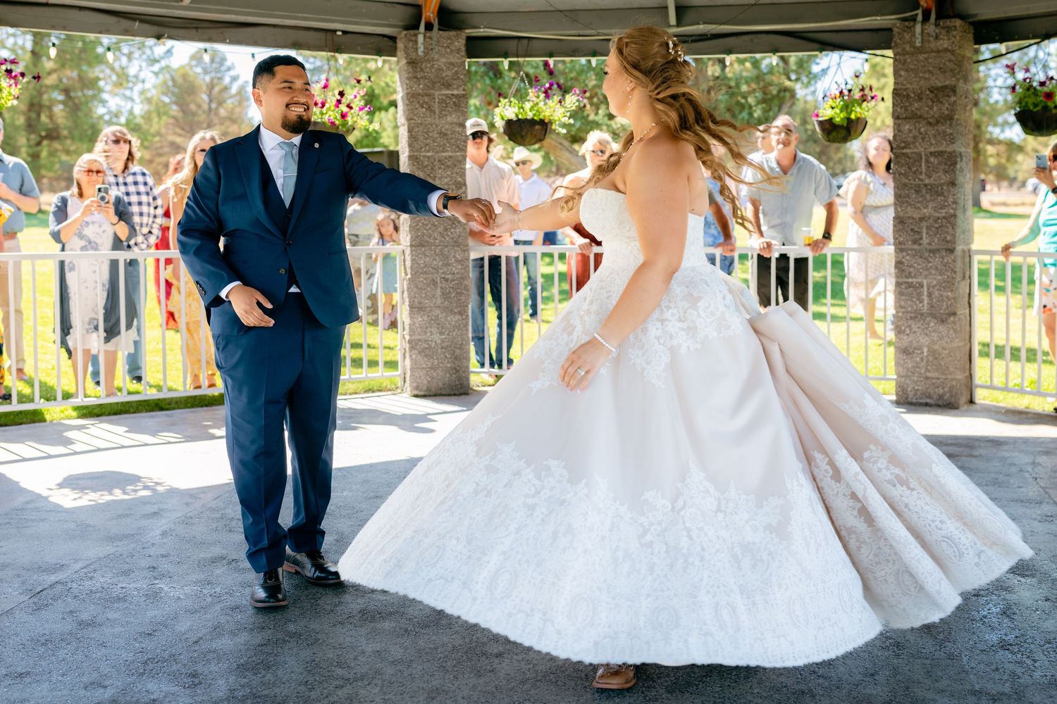 Bride and groom share a joyful first dance in a gazebo surrounded by cheering wedding guests outdoors.