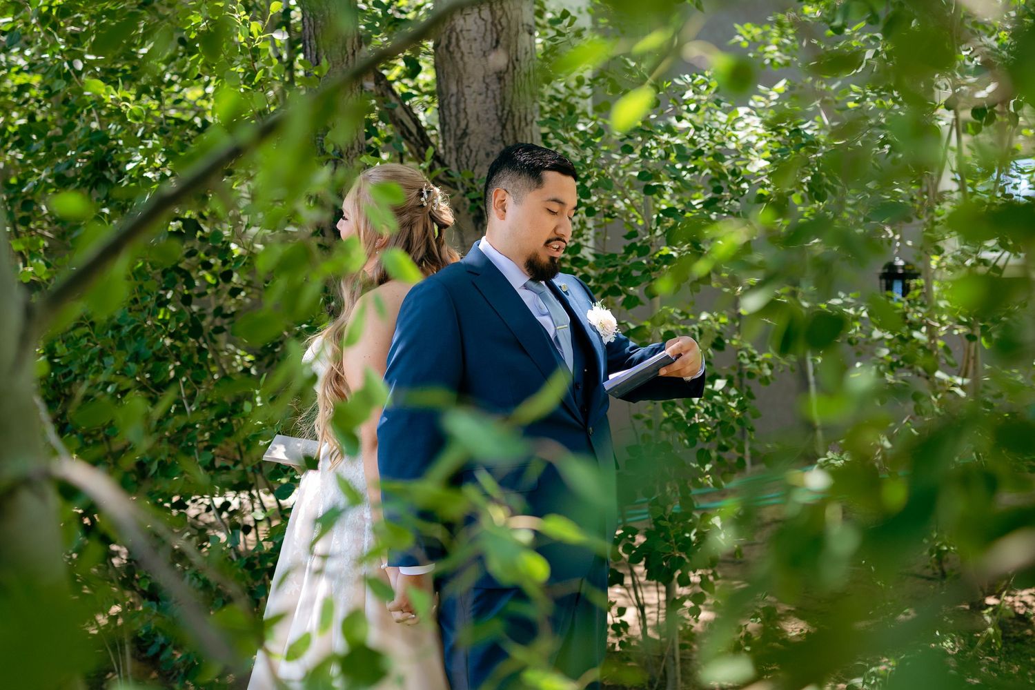 Groom in navy suit reads vows to bride surrounded by lush green foliage during outdoor ceremony.