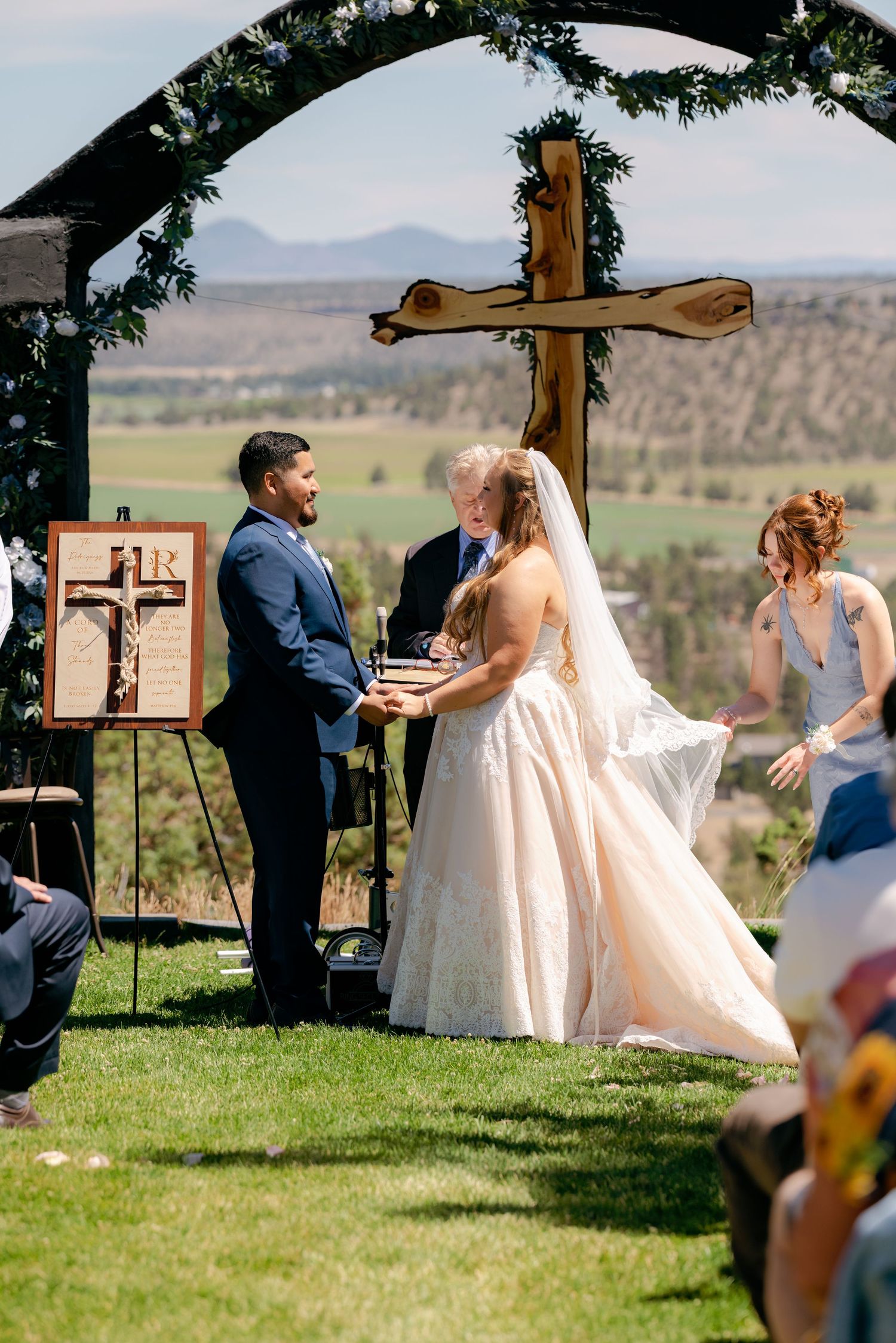 Outdoor wedding ceremony with couple exchanging vows under wooden cross arch with scenic mountain valley backdrop.