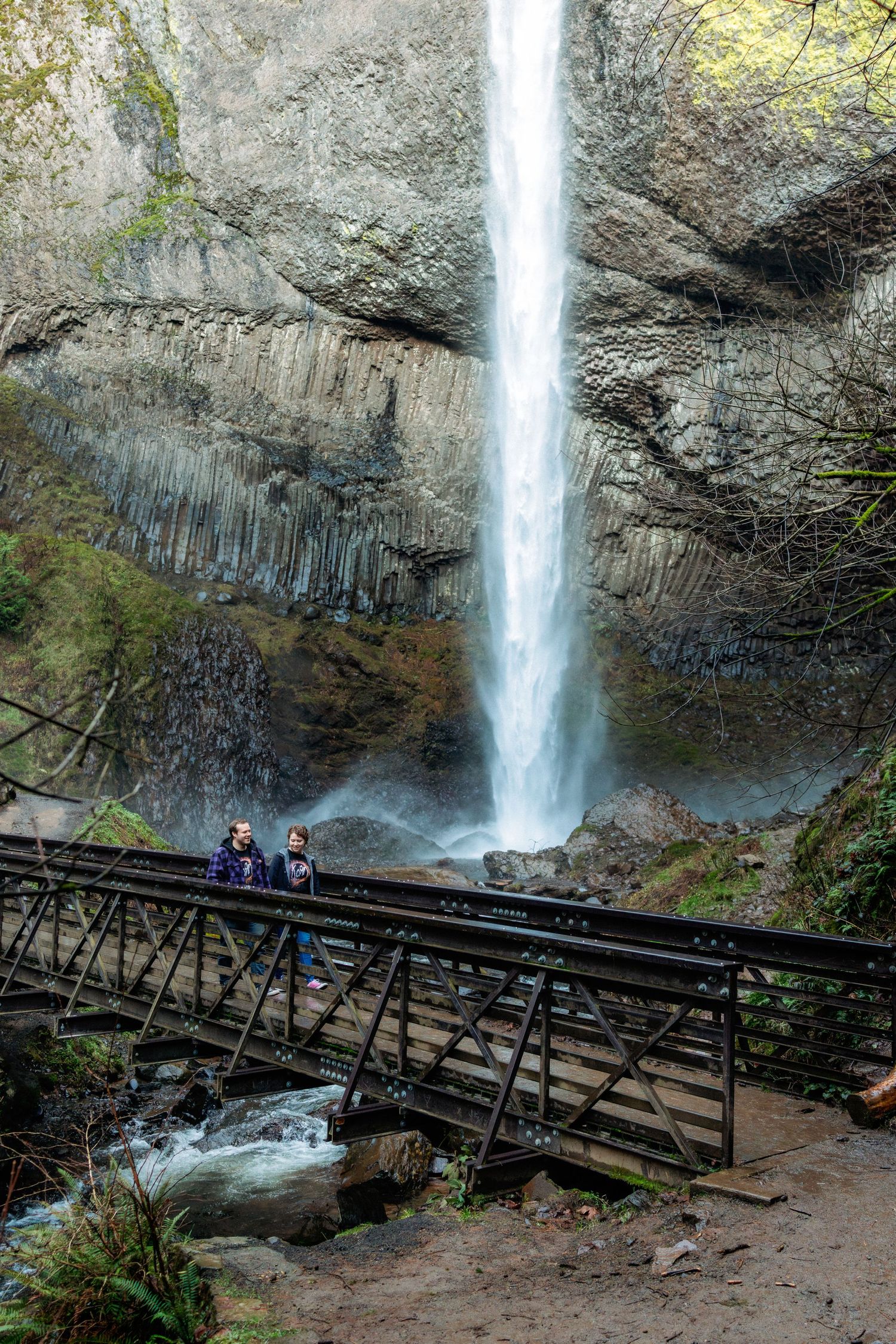Visitors walk along a wooden boardwalk viewing platform beneath a towering waterfall cascading down rocky cliffs.